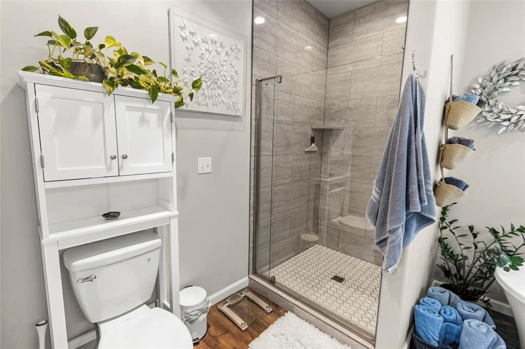 Modern bathroom with a walk-in shower, toilet with a white wood cabinet above, decorative plant on top, wall art, towels hanging, and a potted plant beside a bathtub.