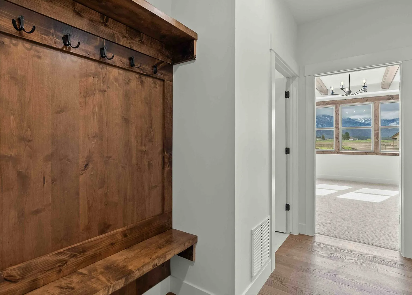 An entryway with a wooden coat rack with hooks on the left and a view into a bright room with large windows showing mountains and blue skies outside.