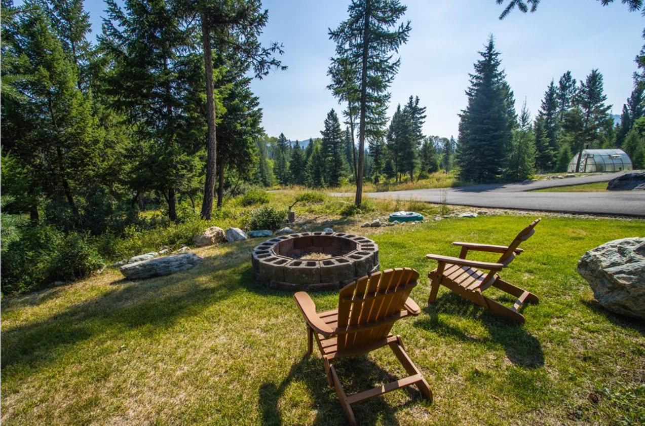 A backyard scene with two wooden chairs around a circular brick fire pit, large rocks, green grass, tall trees, and a road in the background under a clear blue sky.