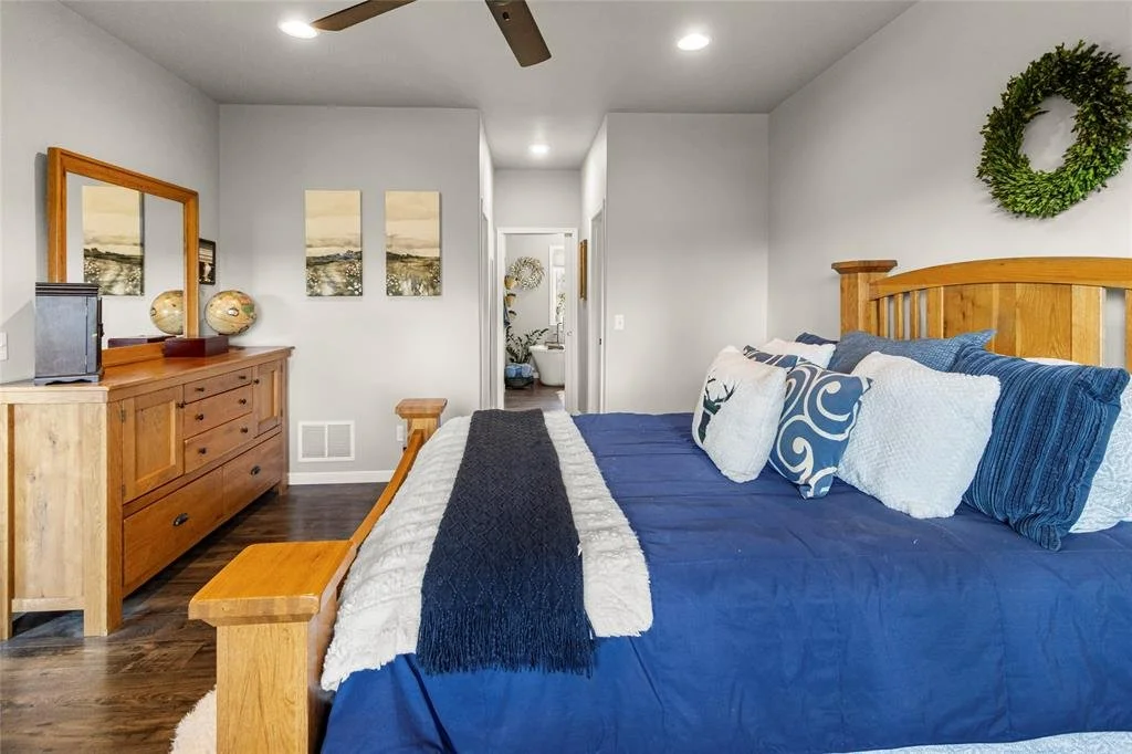 Bedroom with a wooden bed frame, blue bedding, decorative pillows, a wooden dresser with a mirror and decor, a wreath on the wall, and a view into a bathroom.
