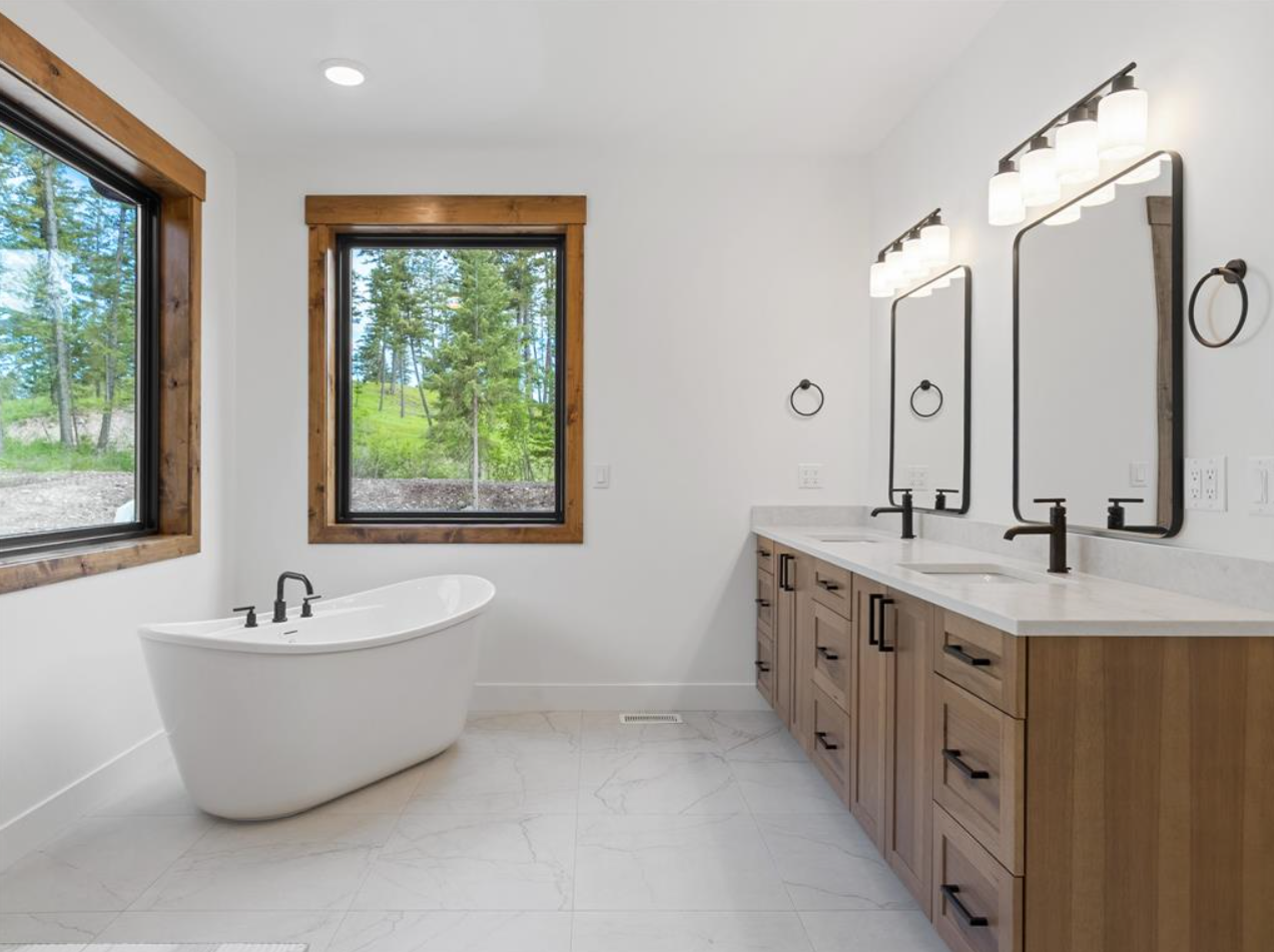 Modern bathroom with white walls, two windows framed in wood, a freestanding bathtub, and a wooden vanity with two sinks and black fixtures, illuminated by wall-mounted light fixtures.