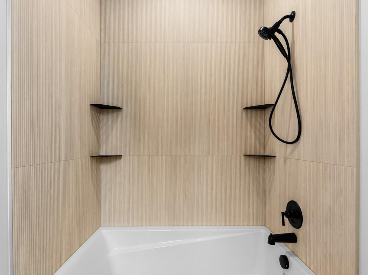 A modern shower with a black handheld showerhead, a black faucet, and two black corner shelves, with light wood-paneled walls and a white bathtub.