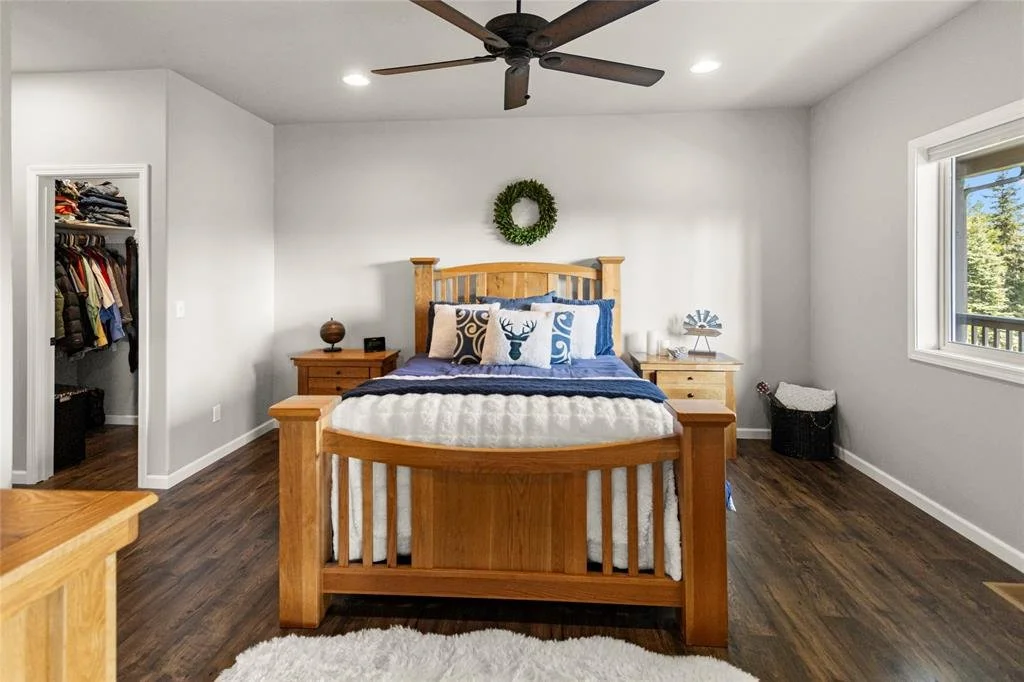 Bedroom with wooden bed frame, white walls, hardwood floors, a window on the right, and a closet on the left. Decor includes a green wreath above the bed and pillows with deer and swirl patterns.