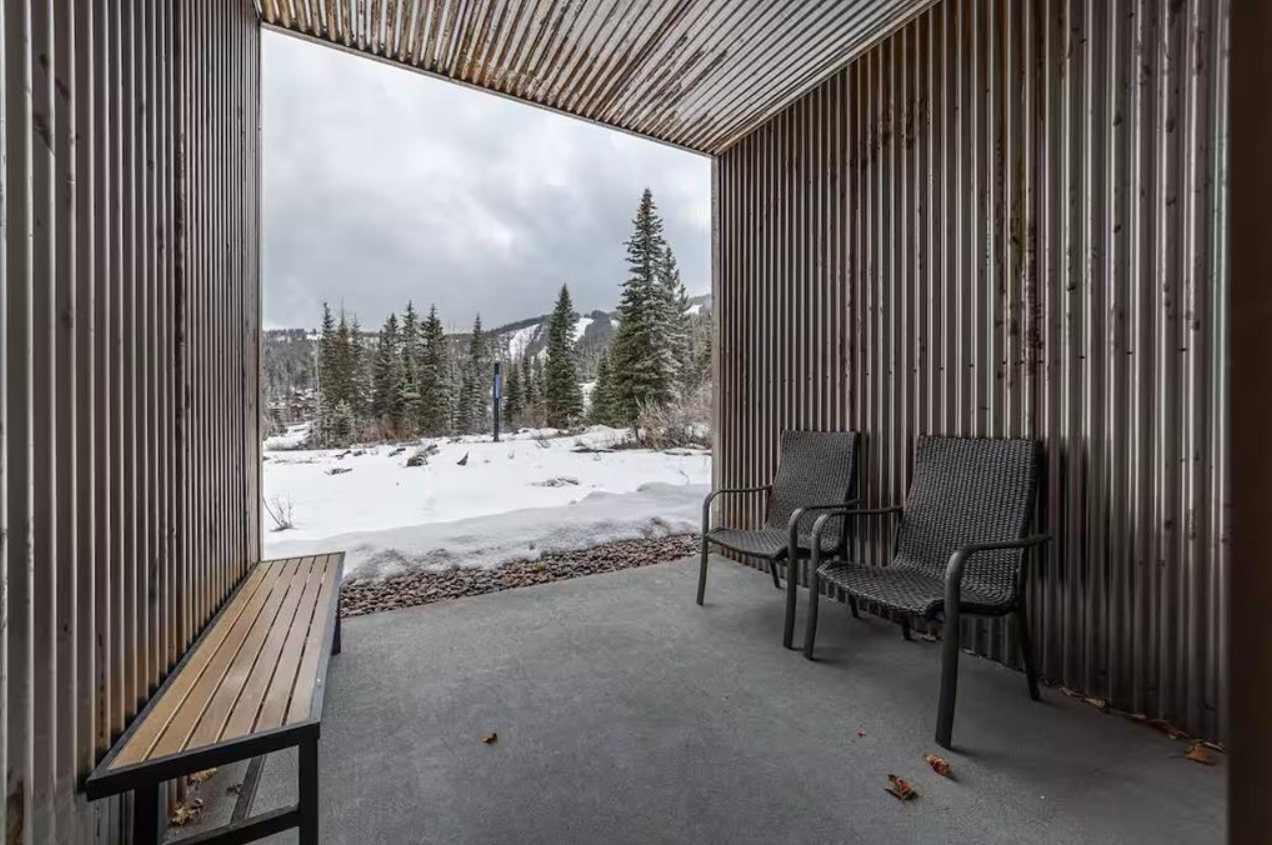 View of a snowy landscape with evergreen trees and a mountain in the background, seen from a covered porch with metal walls and two black woven chairs.