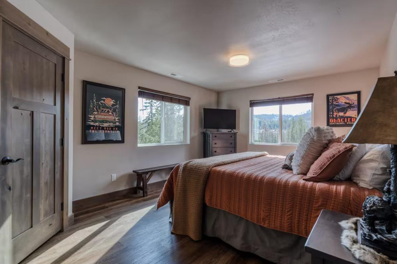 A cozy bedroom with a large bed covered in a rust-colored quilt and decorative pillows. The room features two windows with views of a forested landscape, a flat-screen TV on a wooden dresser, artwork on the walls, and wooden flooring with a bench ben