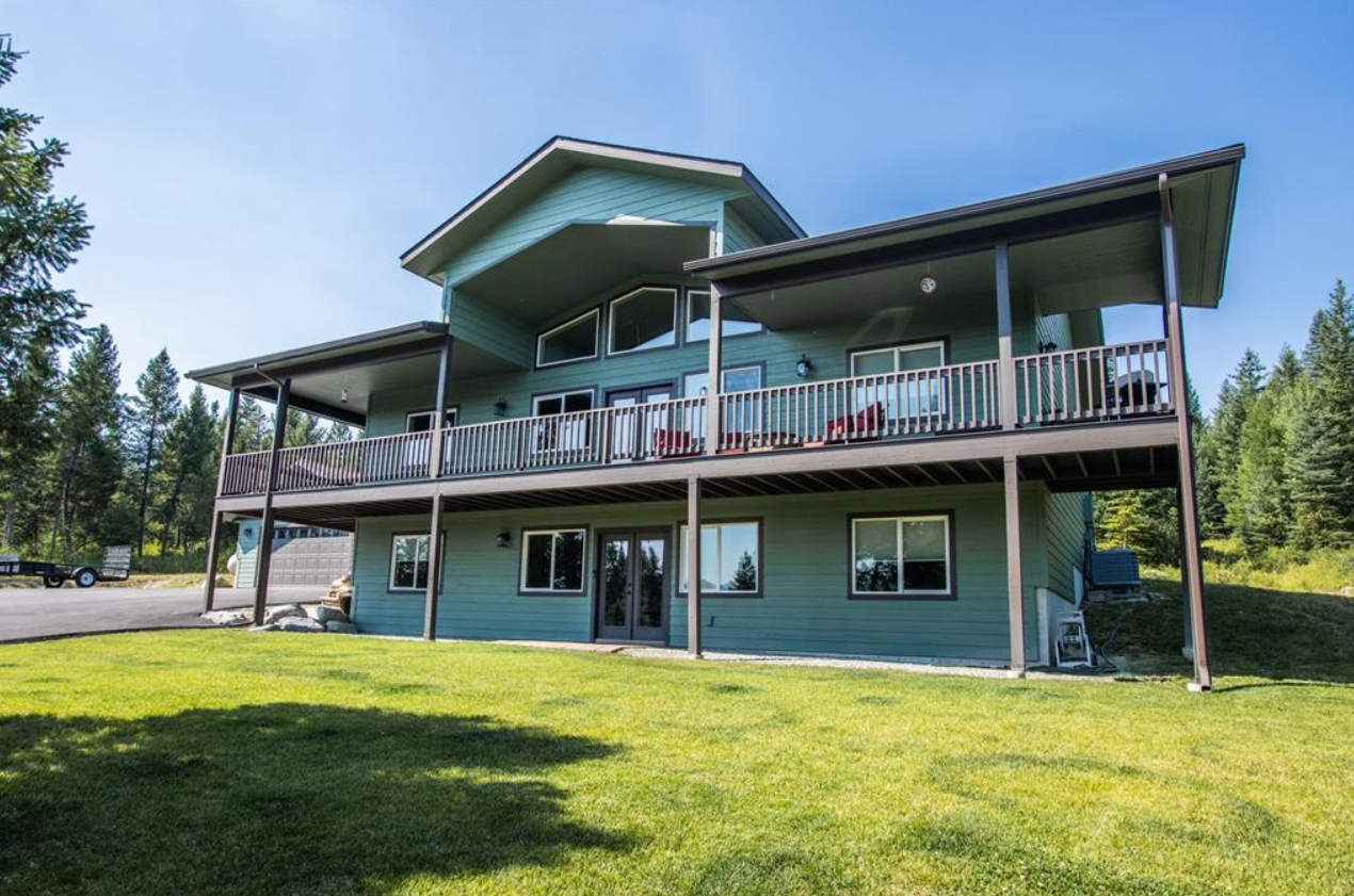 A large, modern two-story house with a green exterior and a spacious upper deck, located in a lush, wooded area with a well-maintained lawn.