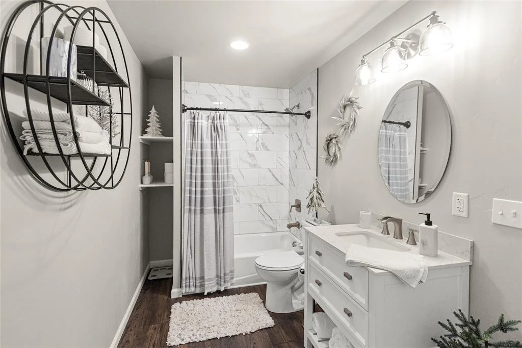 A modern bathroom with a white vanity, round mirror, wall-mounted light fixtures, a bathtub with a shower curtain, and decorative wreaths on the wall.