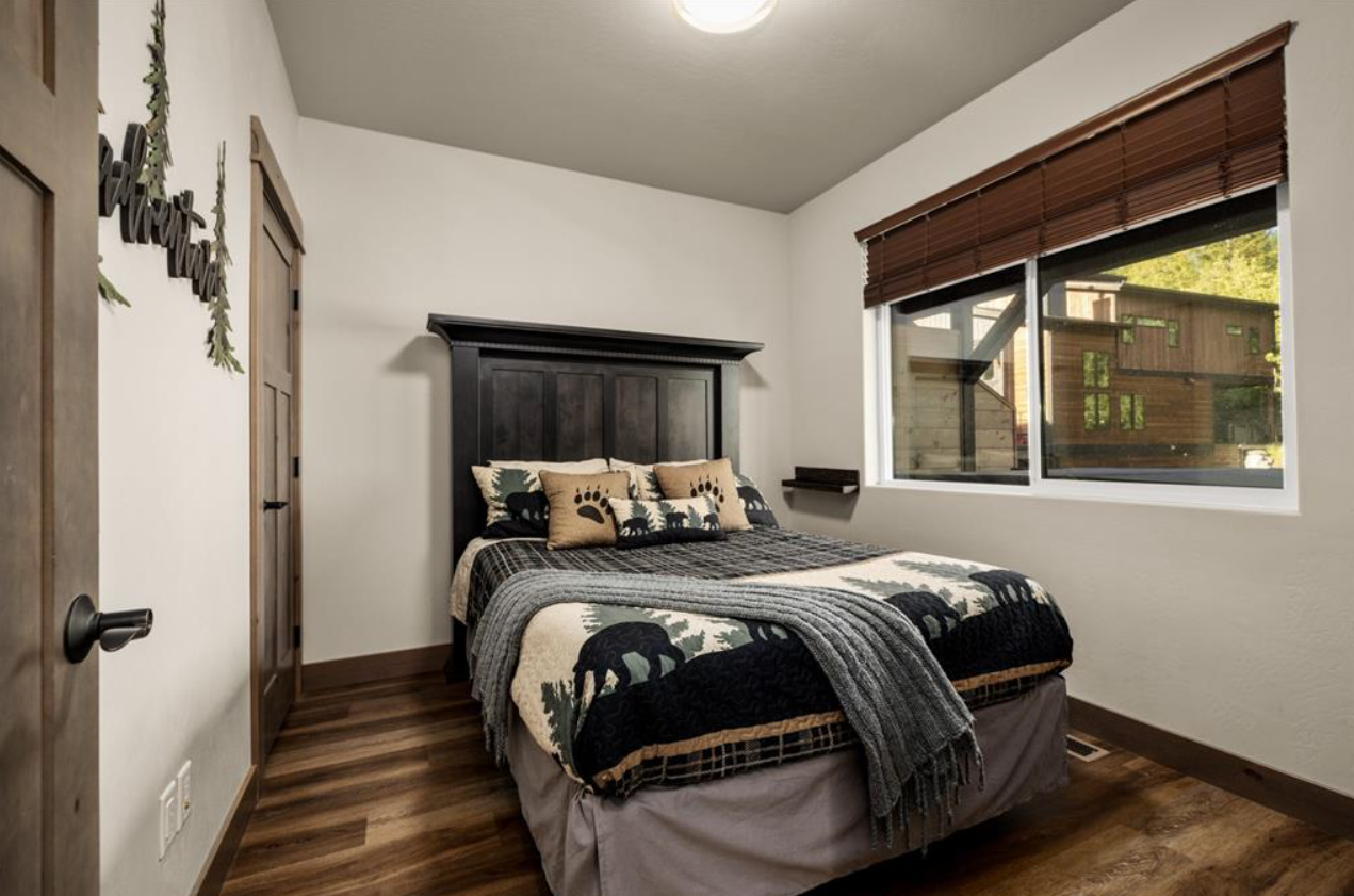 A bedroom with a bed featuring bear and forest-themed pillows and a quilt. There is a window with wooden blinds and a simple ceiling light. The room has white walls, wood flooring, and wooden trim.