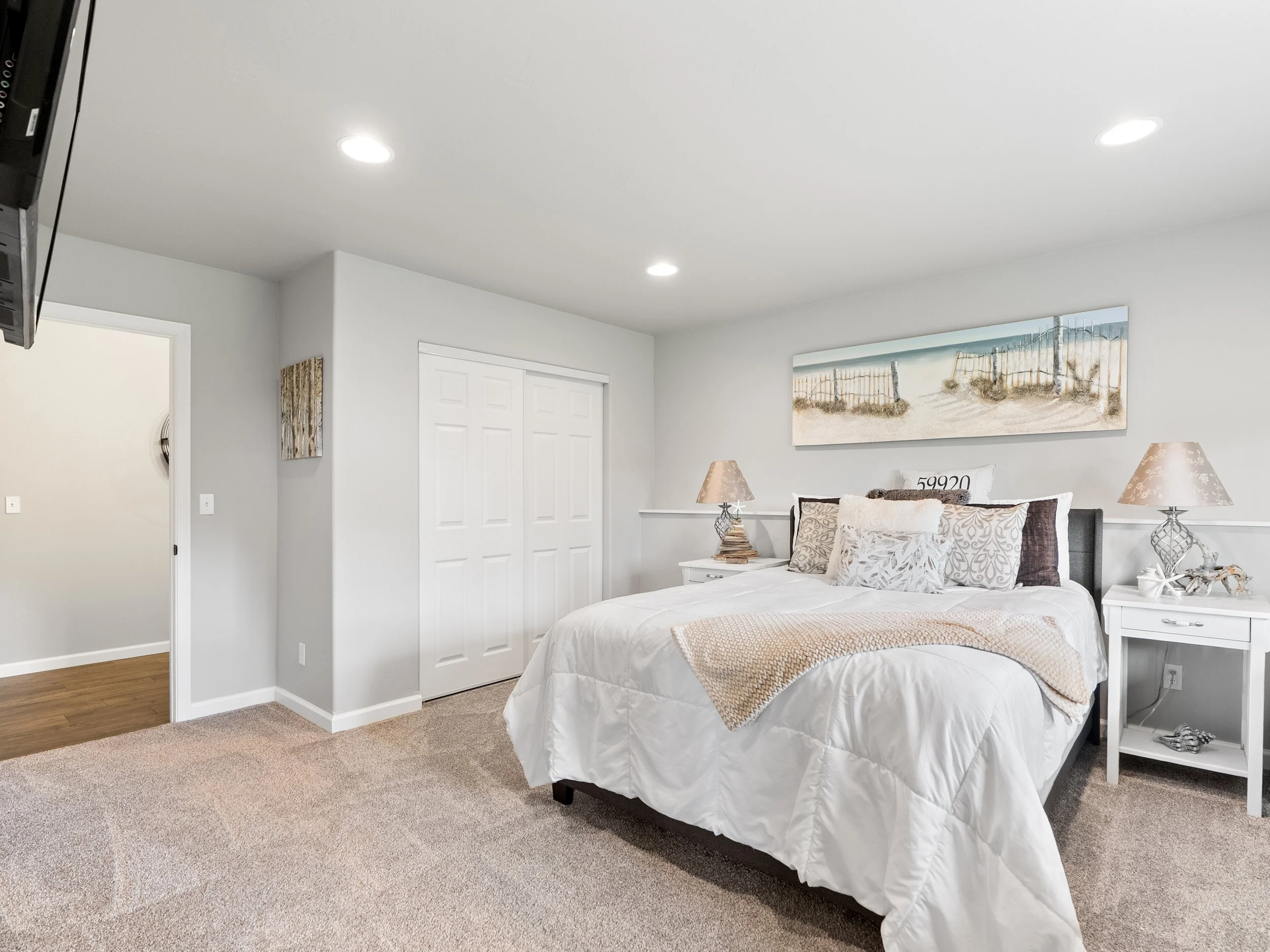 A bedroom with a white bedspread, two table lamps on bedside tables, a wall art of a beach scene, an open door, and a closet.