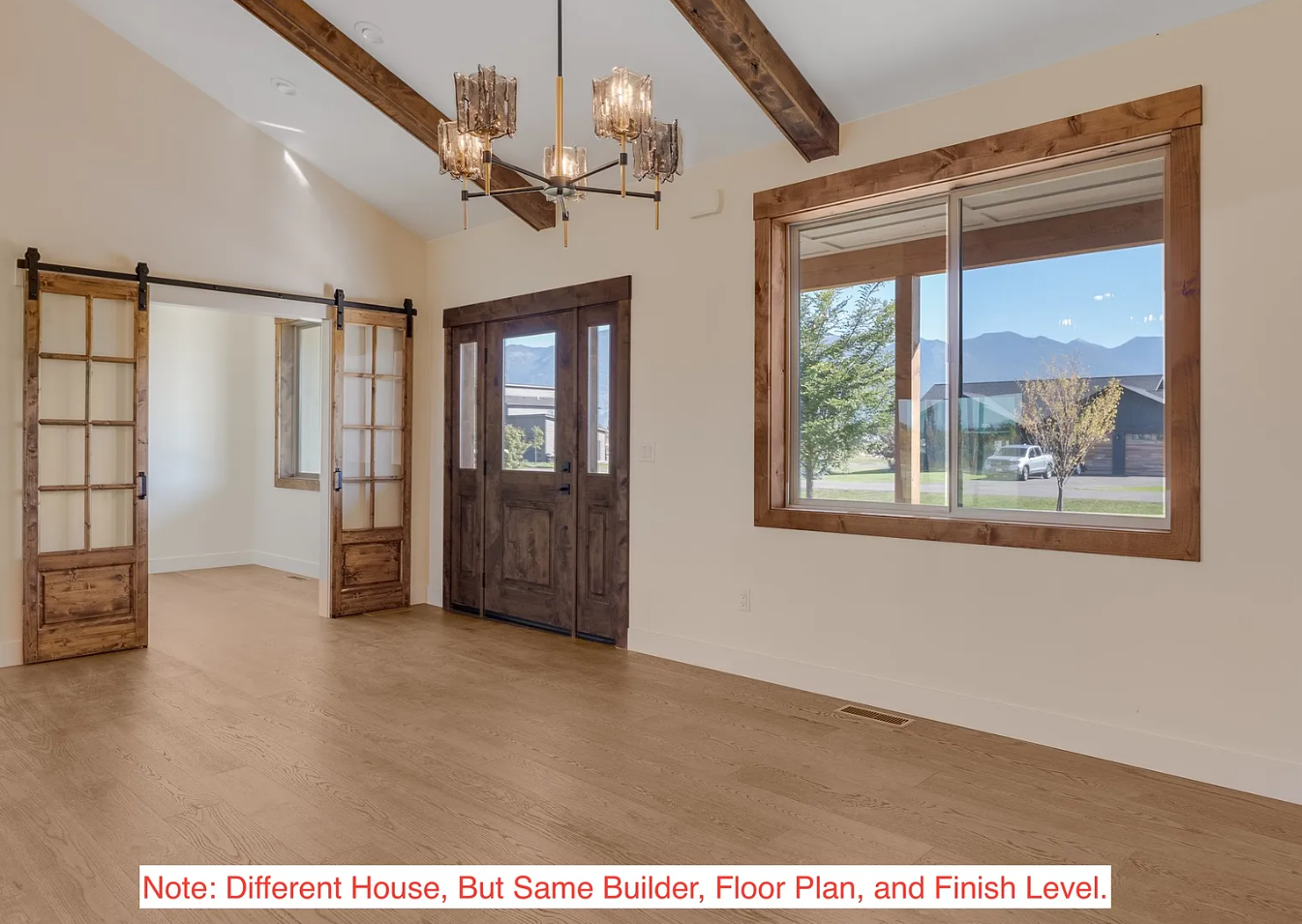 Empty room with wooden floors, large window showing outdoors, dark wooden door, sliding doors with wooden frames, wooden ceiling beams, and a vintage chandelier.