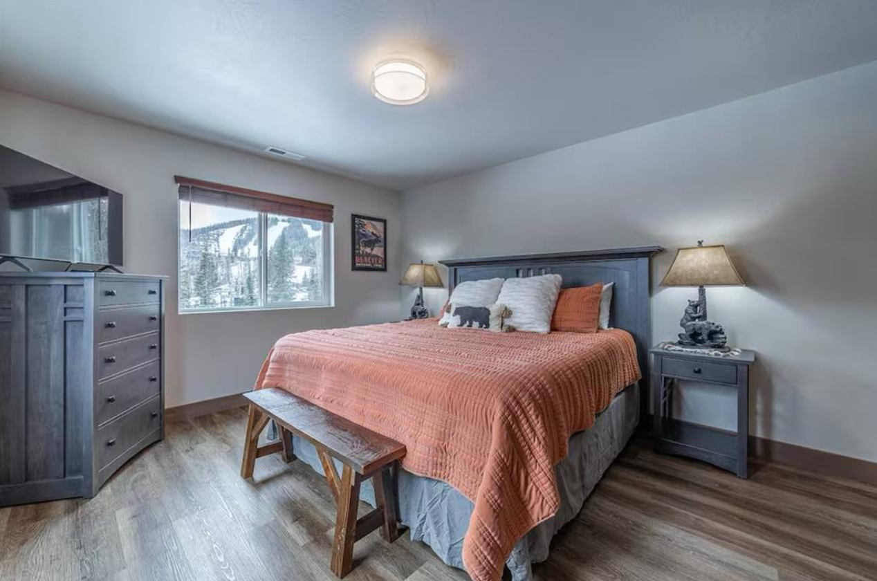 A bedroom with a large bed covered with an orange quilt, decorative pillows, two bedside tables with lamps, a wooden bench at the foot of the bed, a dresser, a colorful framed picture on the wall, and a window showing a snowy outdoor scene.