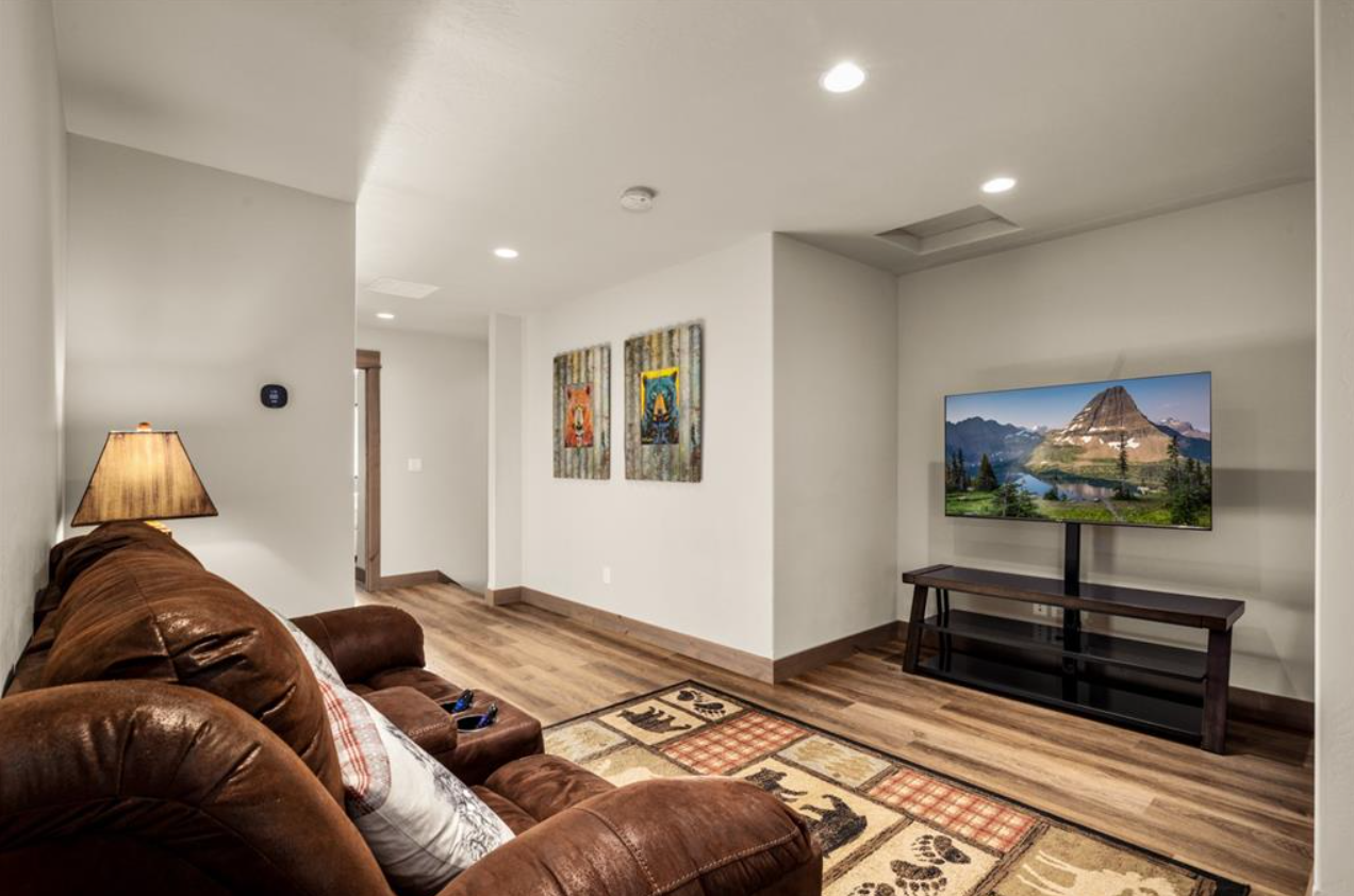 Living room with a brown leather sofa, a side table with a lamp, wall-mounted TV displaying a mountain landscape, hardwood floors, wall art, and a colorful area rug.