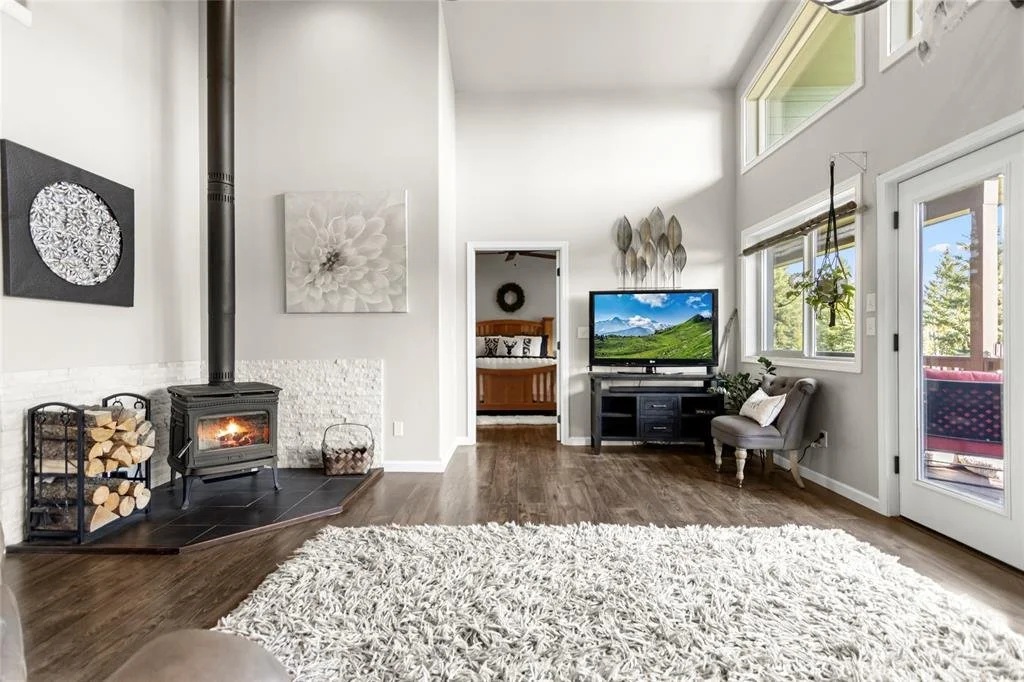 Living room with wood stove, stacked firewood, white walls, large window, TV on black stand, and doorway leading to another room.