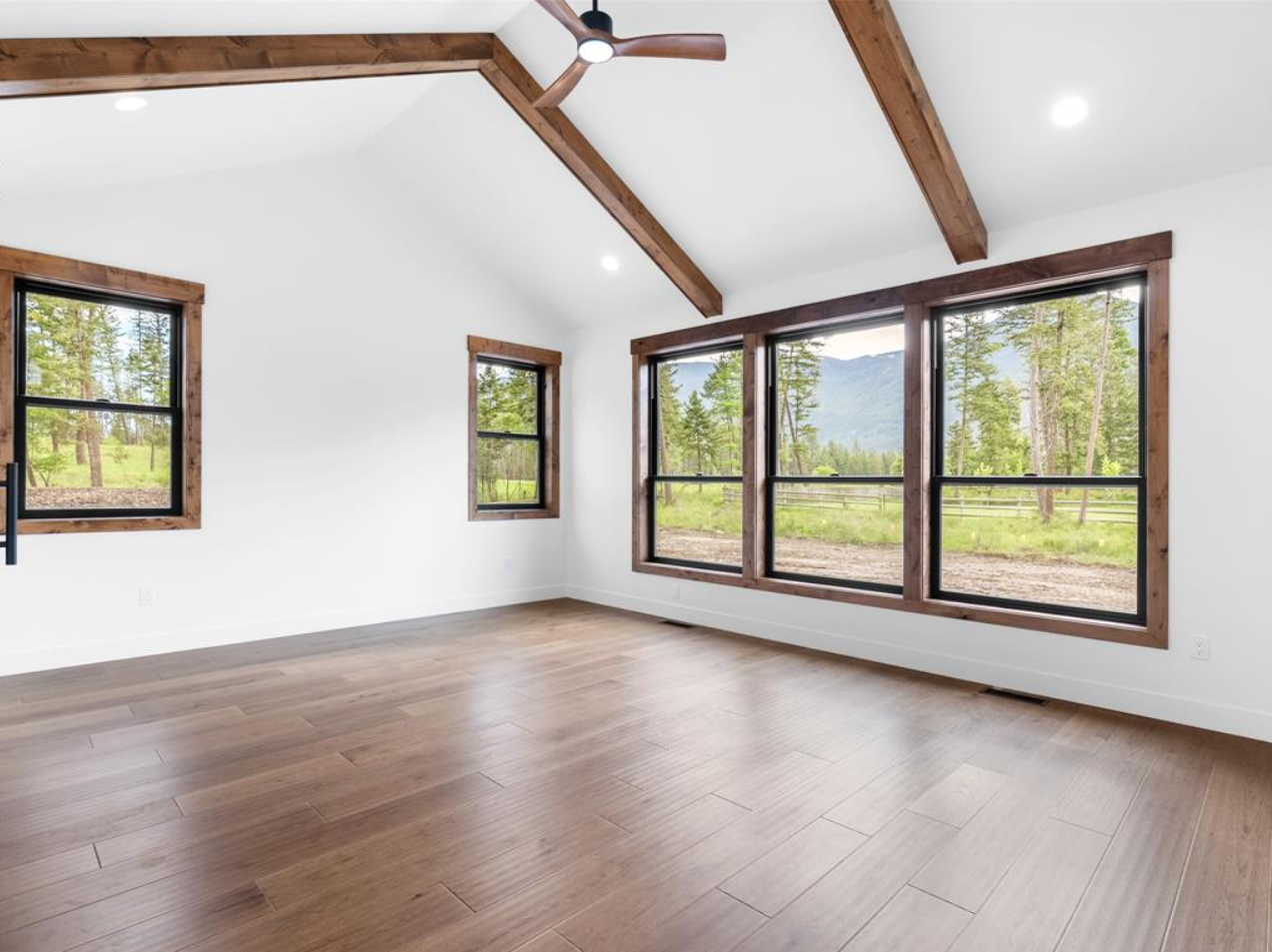 Empty room with large windows, wooden beams, and wood flooring, overlooking a scenic outdoor landscape with trees and mountains.