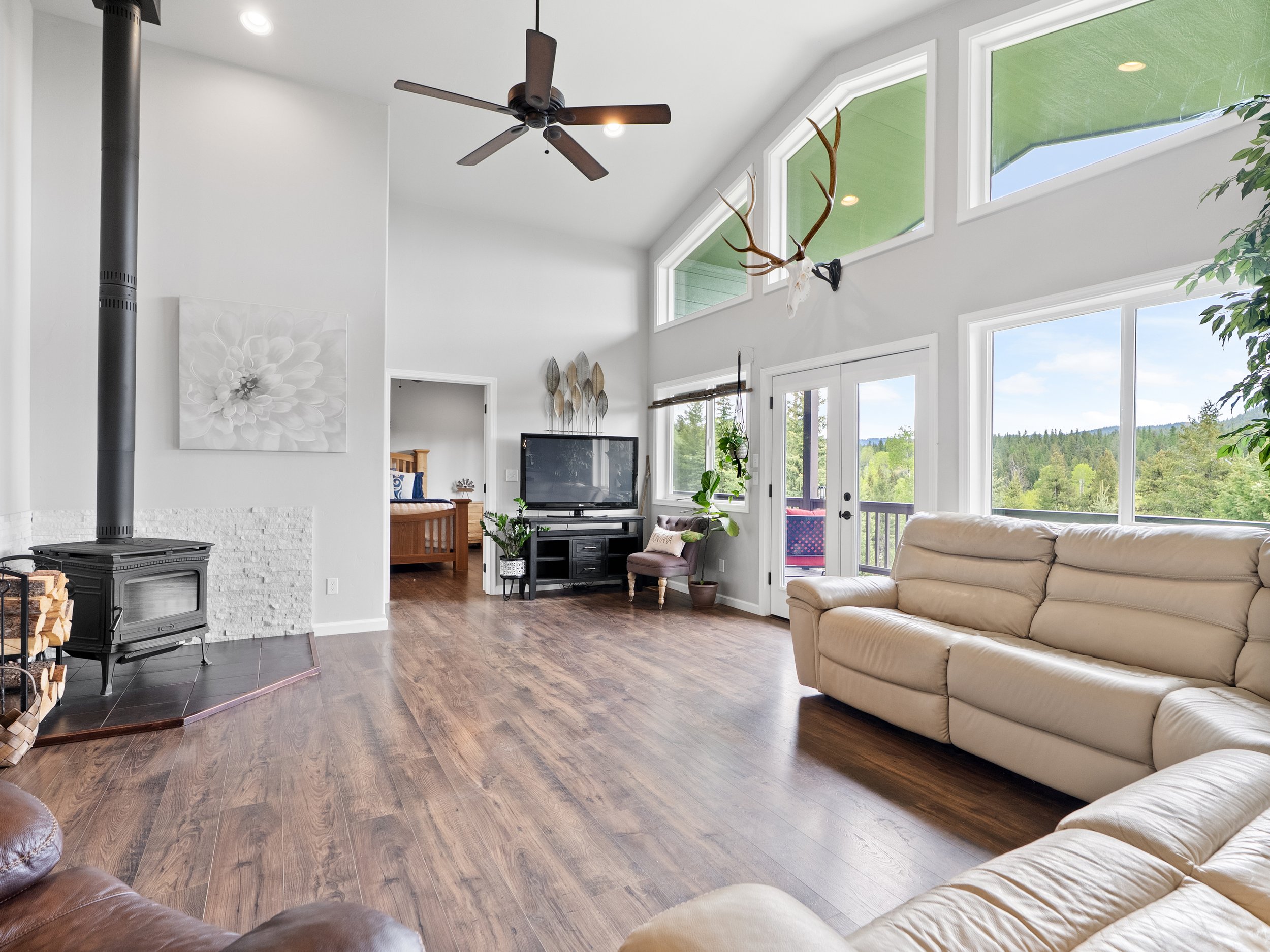A spacious living room with large windows showing a view of trees and a blue sky, a cream-colored sofa, a black television, a fireplace with a wood stove, plants, and a ceiling fan.