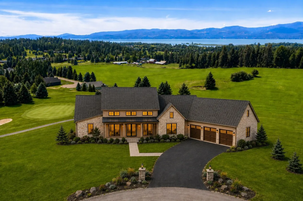 A large modern house with a dark roof and white exterior walls, surrounded by a well-maintained lawn and landscaping, with a curved driveway leading to a three-car garage, set against a scenic landscape of green fields, trees, mountains, and a body of water in the background.