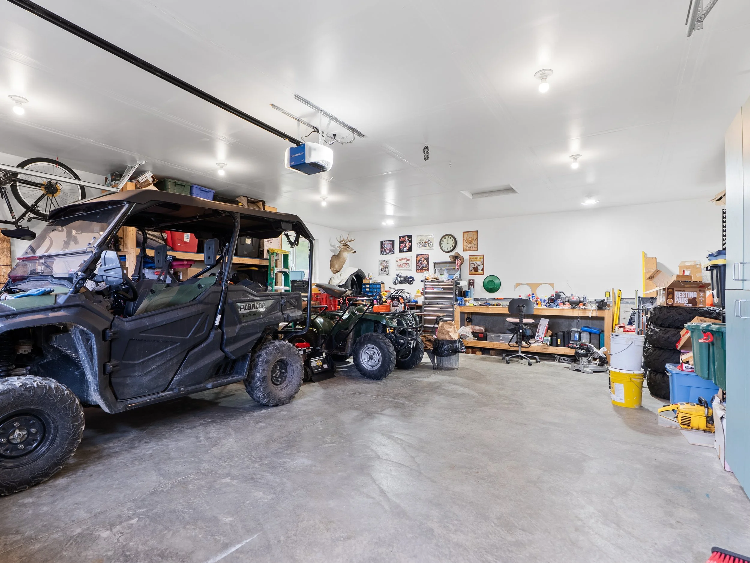 Inside a spacious garage with a white ceiling, fluorescent lights, and concrete floor. Several off-road utility vehicles are parked, with tools, shelves, and storage boxes along the walls. The garage includes decorations such as mounted animal heads,