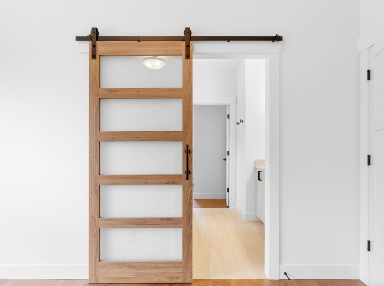 Wooden sliding barn door on black metal track in a white room, partially open to reveal a hallway with a light-colored floor and white walls.