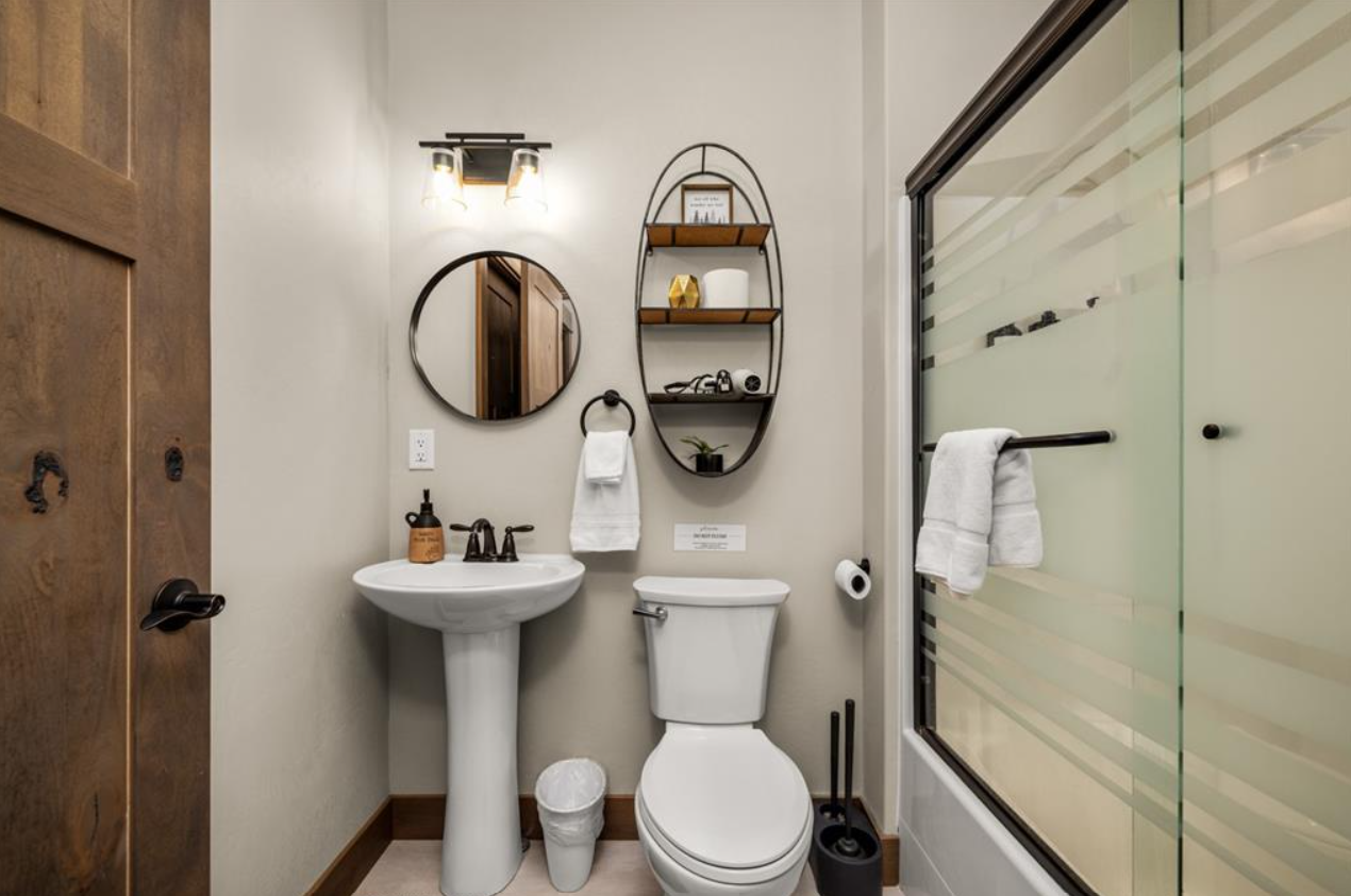 A small modern bathroom with a white pedestal sink, round mirror, black fixtures, a white toilet, and a glass shower door with a towel hanging on a black towel bar. Decorative shelves and a mirror are also visible.