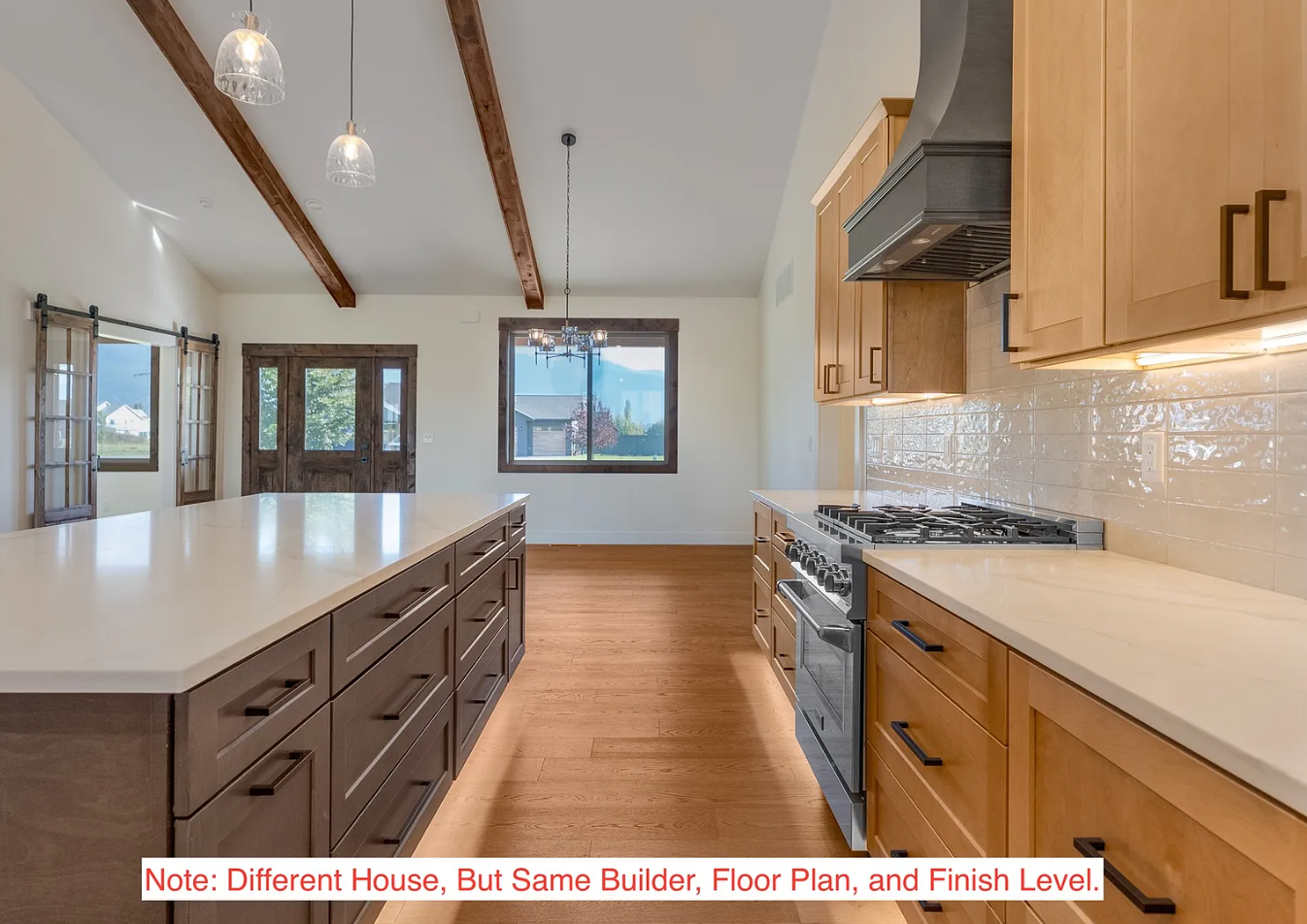 Interior view of a modern kitchen with wooden cabinets, a gas stove, a large island with drawers, hardwood flooring, windows, and a sliding door, with ceiling exposed beams and hanging pendant lights.