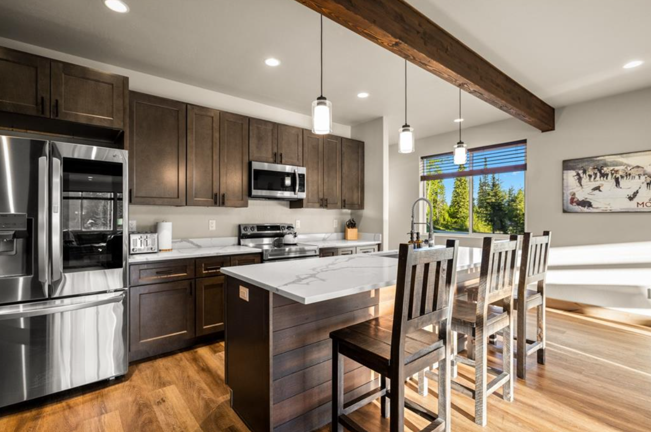 Modern kitchen with dark wood cabinets, stainless steel refrigerator and microwave, white marble countertop island with sink, three wooden barstools, large window with blinds, and wood beam ceiling.