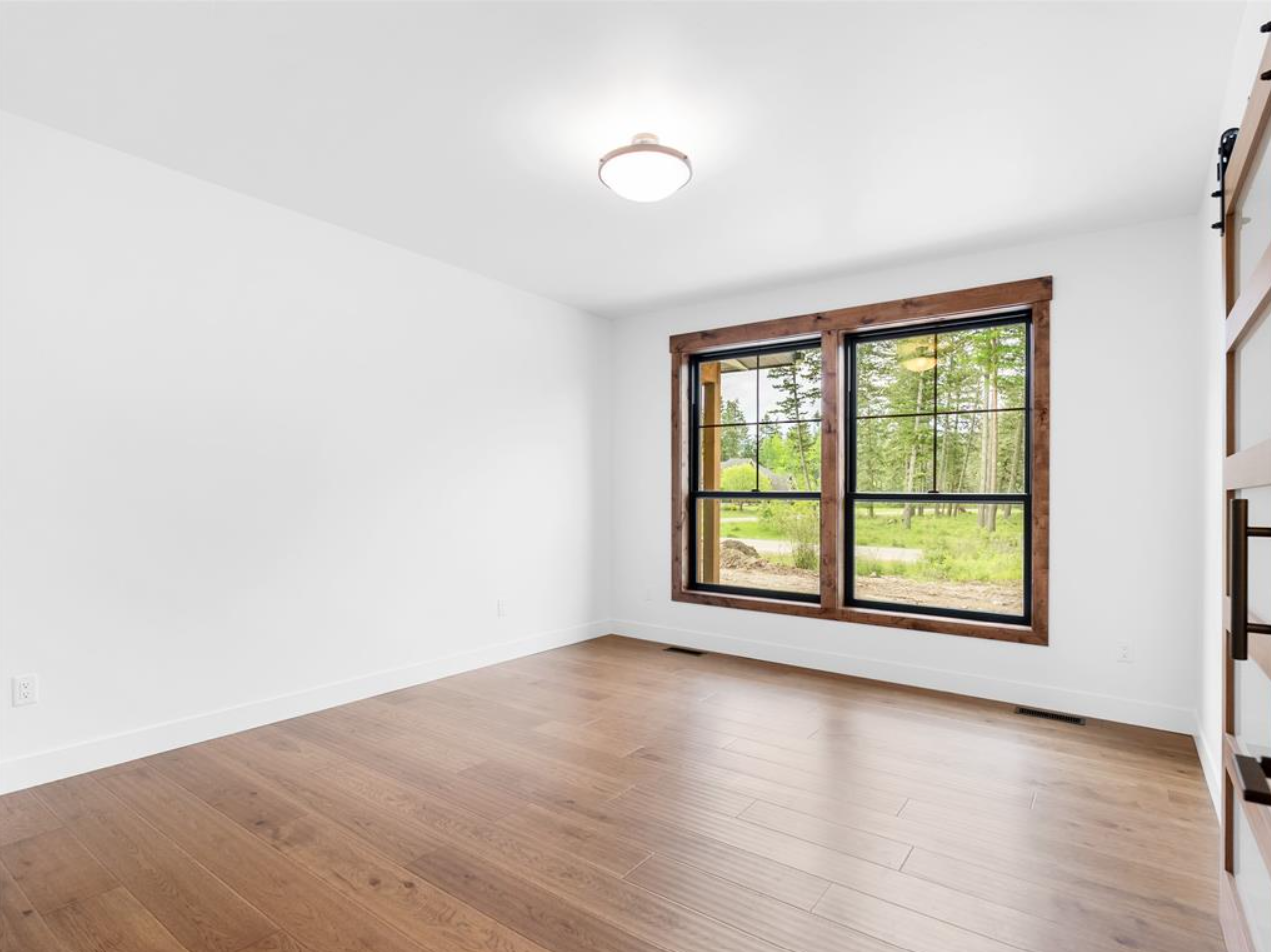 Empty room with hardwood floors, white walls, large window with wooden frame, and a ceiling light fixture, overlooking a green outdoor landscape.
