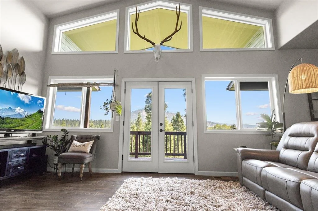 Living room with large windows, a flat-screen TV, a cushioned armchair, a leather sofa, a hanging lamp, and a deer skull mounted above the windows.
