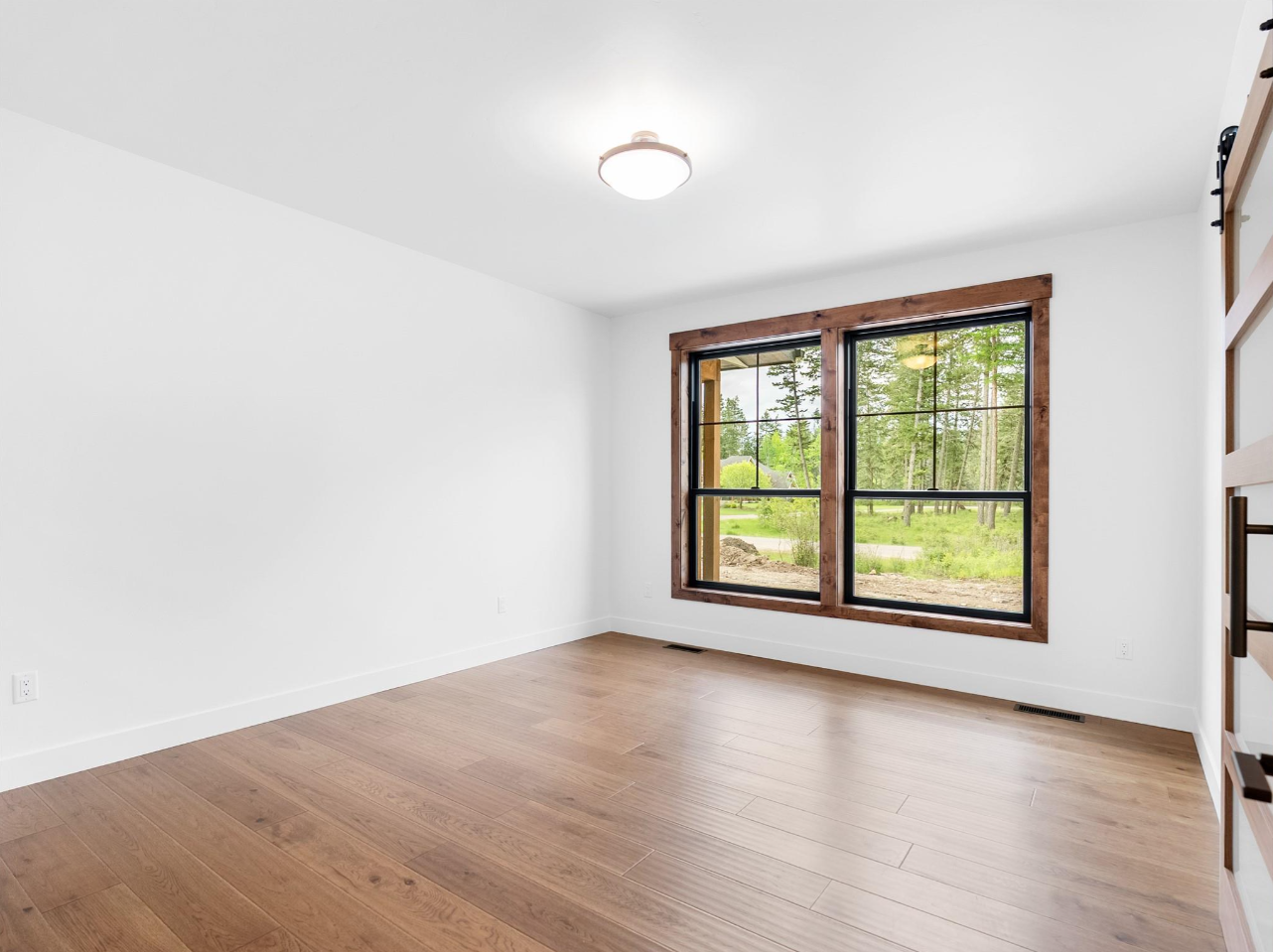 Empty room with wood flooring, large window with wooden frame, and white walls, overlooking a green outdoor landscape.