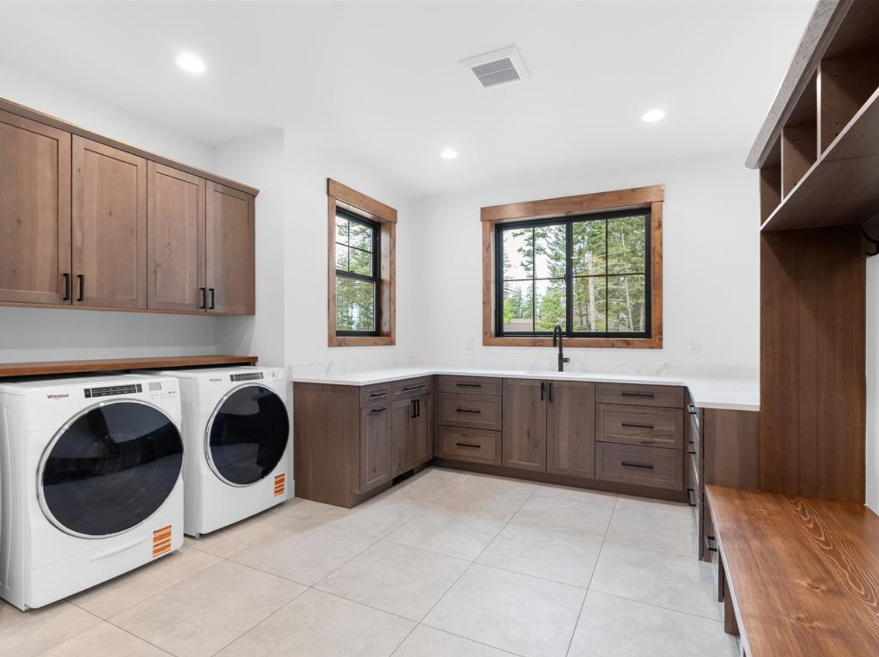 Laundry room with white walls, beige tiled floor, wooden cabinets, a white washer and dryer, two windows with wooden frames, and a white countertop.