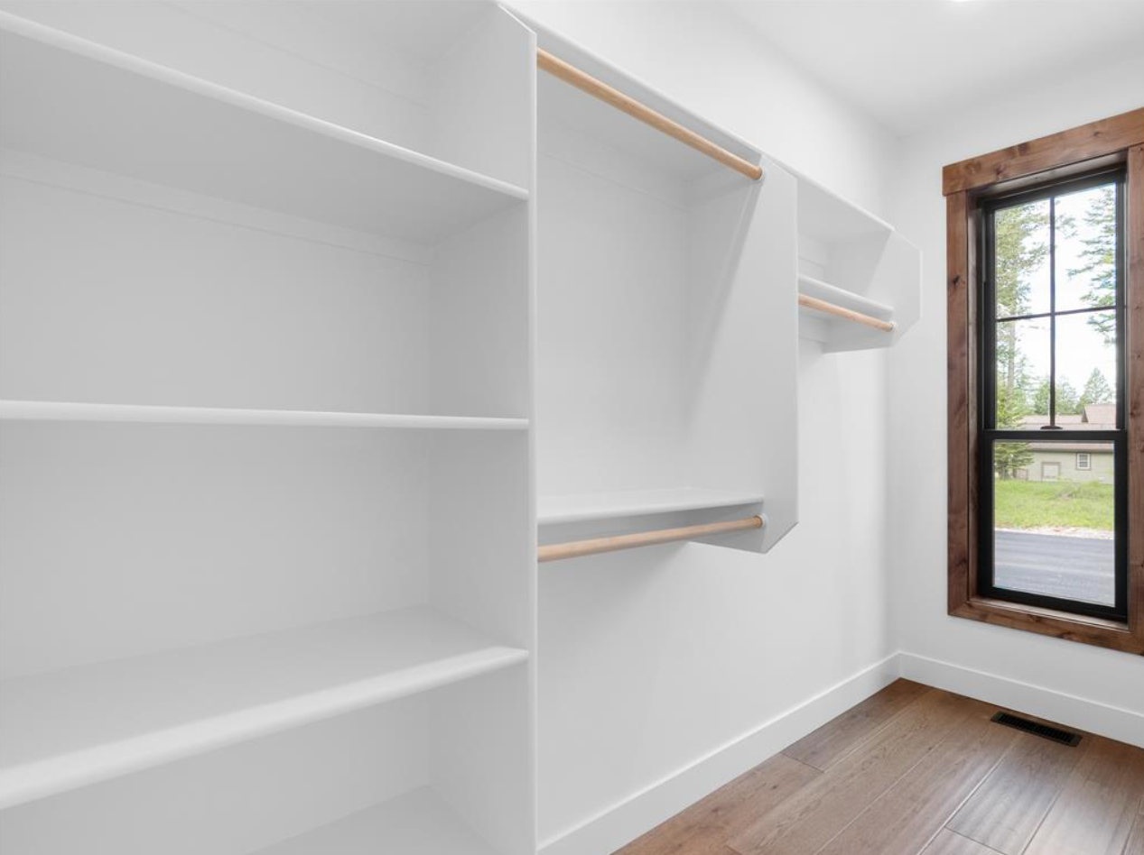 Empty built-in white closet with shelves and hanging rods near a window with wooden trim and a view of trees outside.