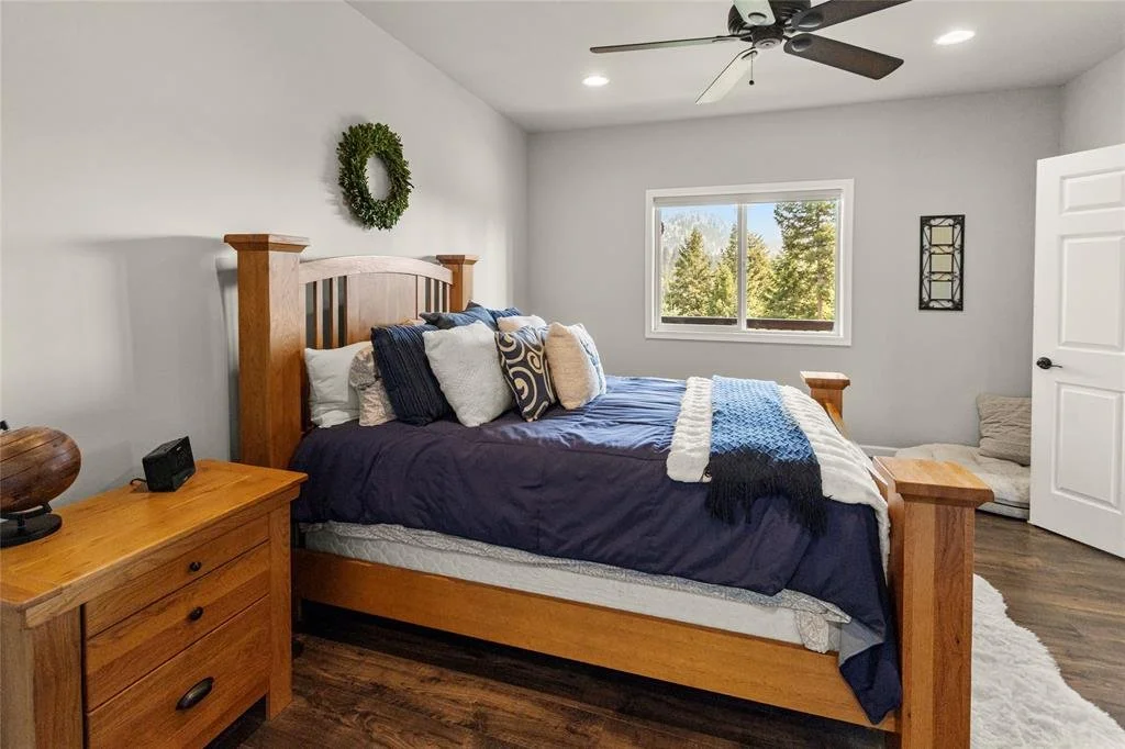 Bedroom with a wooden bed frame, navy blue bedding, decorative pillows, a window showing trees outside, a ceiling fan, and a small sitting area in the corner.