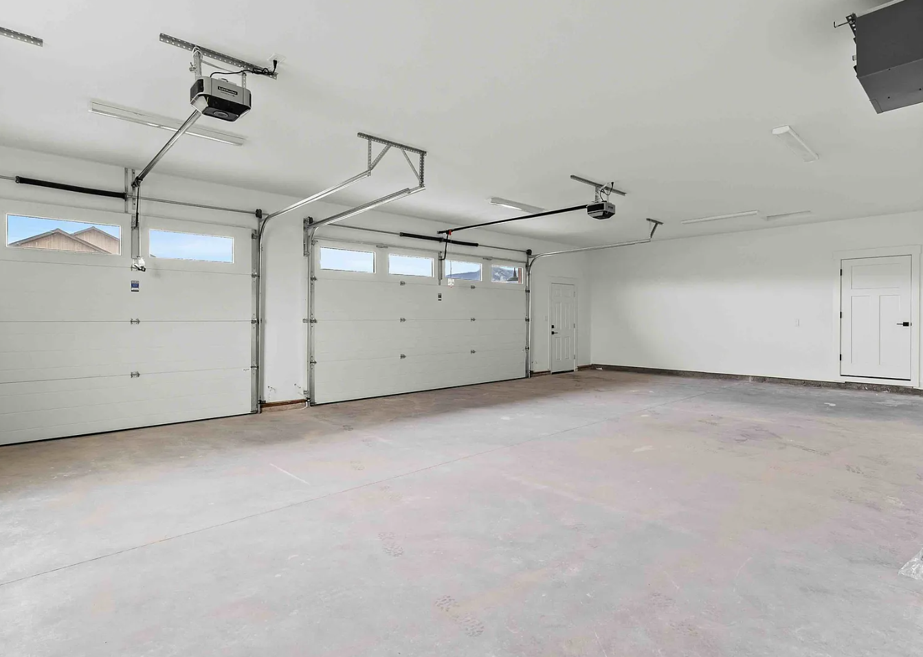 Empty clean two-car garage with white walls, concrete floor, garage doors, small windows, and ceiling-mounted garage door openers.