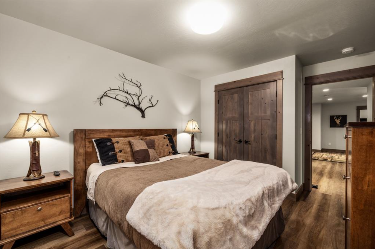 Cozy bedroom with wooden furniture, beige bedding, pillows, bedside lamps, and a decorative branch on the wall, hardwood floor, open closet, and doorway to another room.