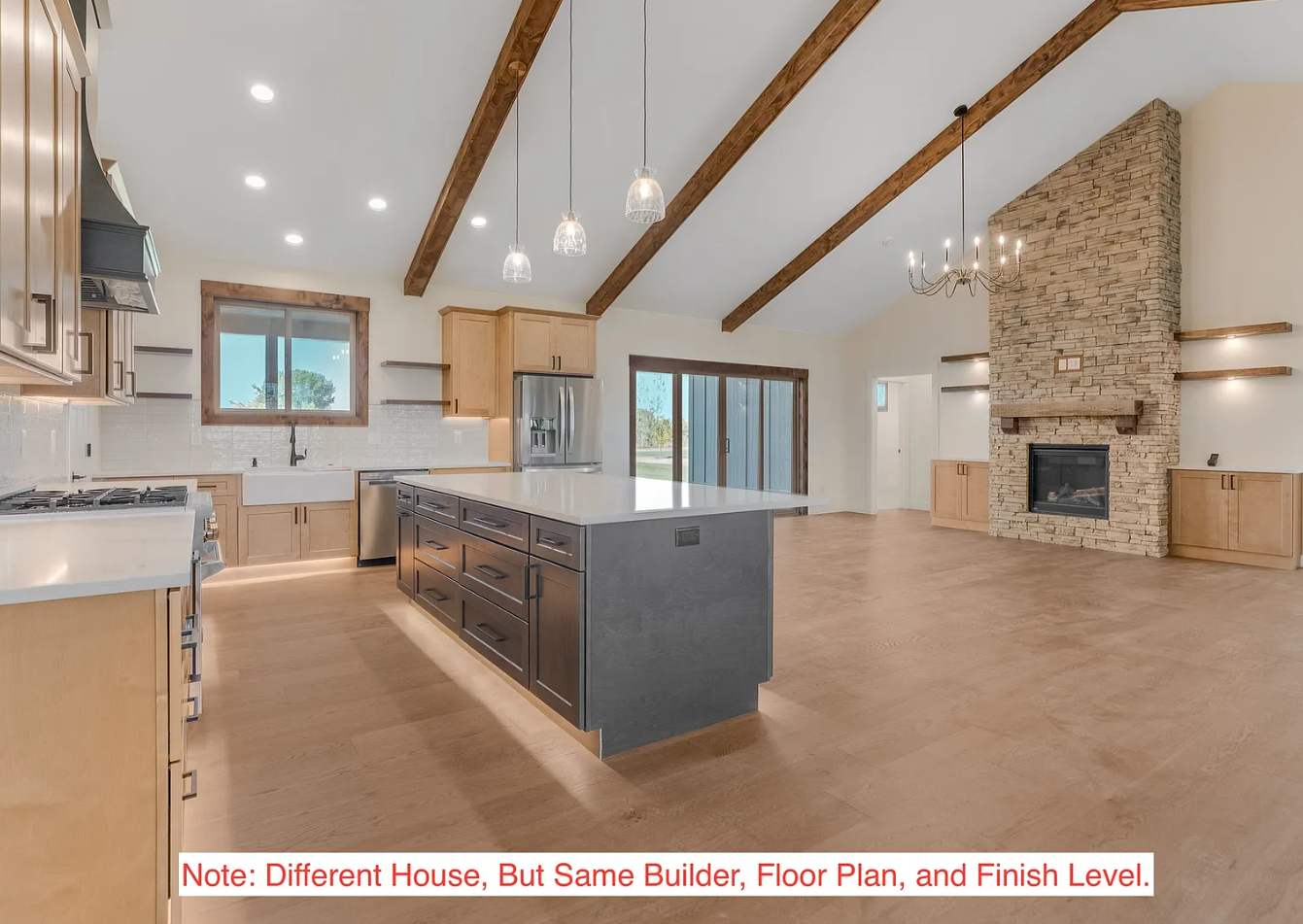 Spacious open-concept kitchen and living room with vaulted ceiling, exposed wooden beams, a stone fireplace, large windows, built-in cabinets, and modern kitchen island.