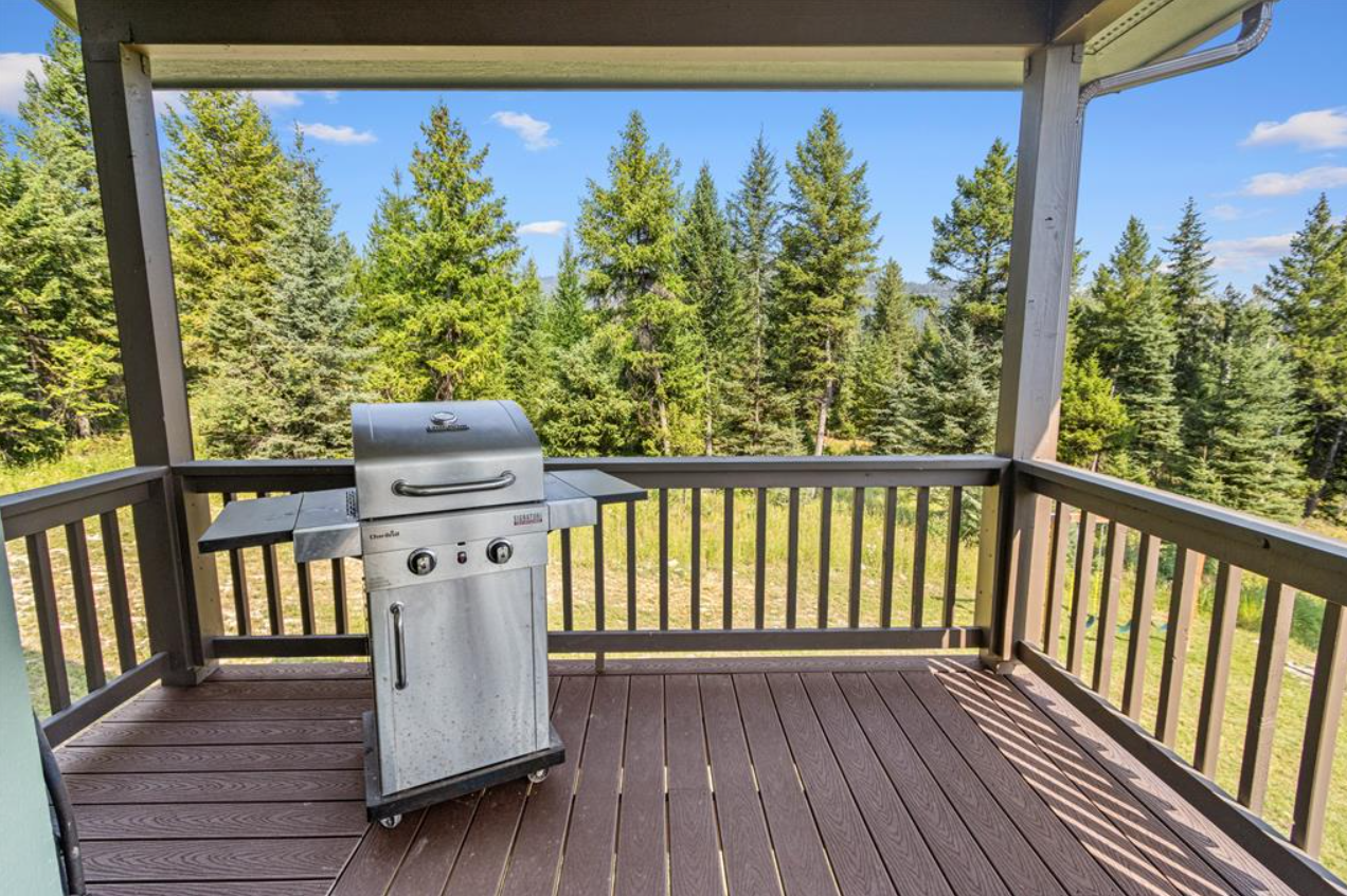 A balcony with a stainless steel grill on a wooden deck, overlooking a forest of green trees under a partly cloudy blue sky.