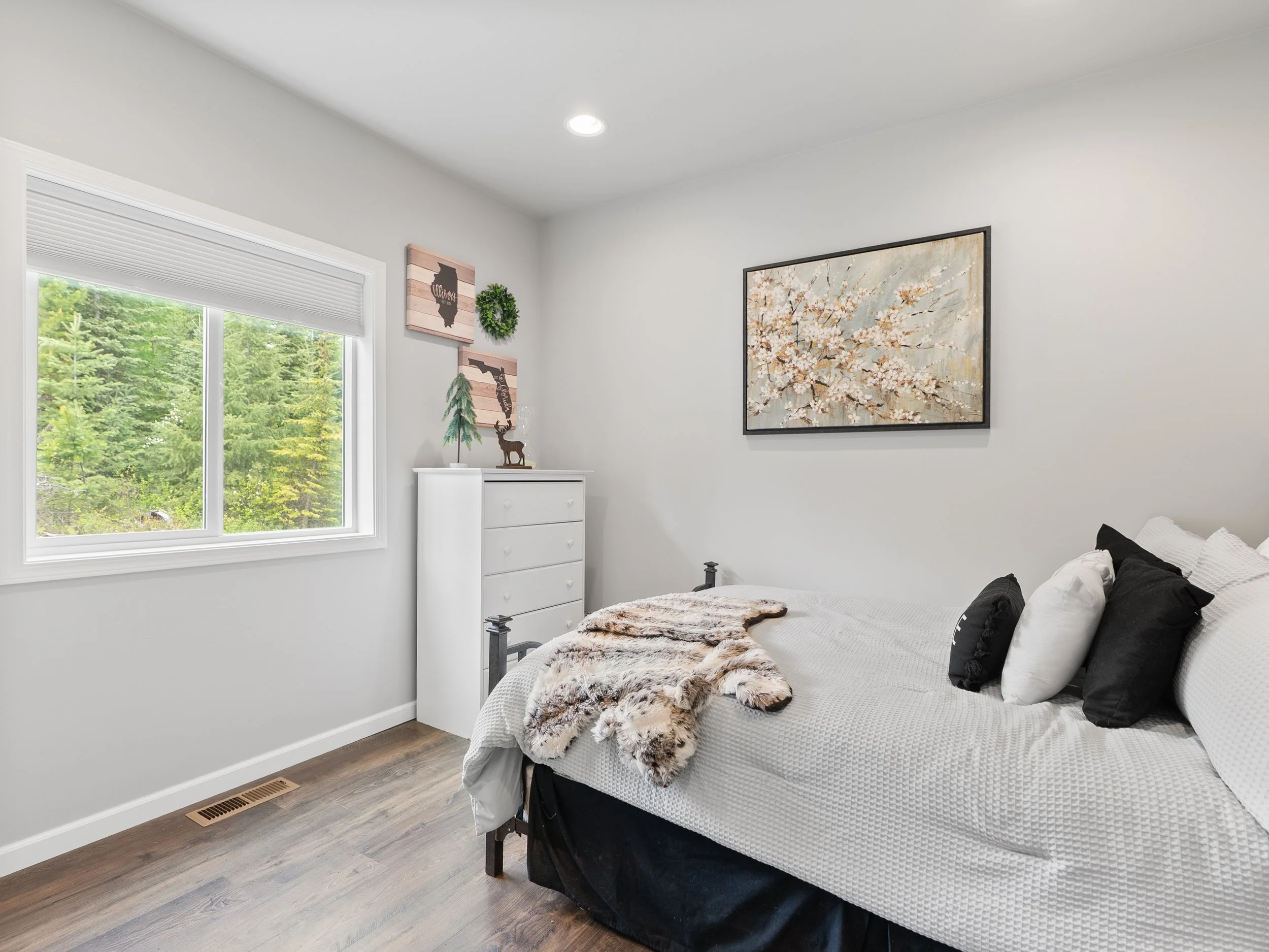 A bright bedroom featuring a window with a view of green trees, a white dresser, wall decorations including artwork and a wreath, a bed with white and black bedding, and a faux fur throw.