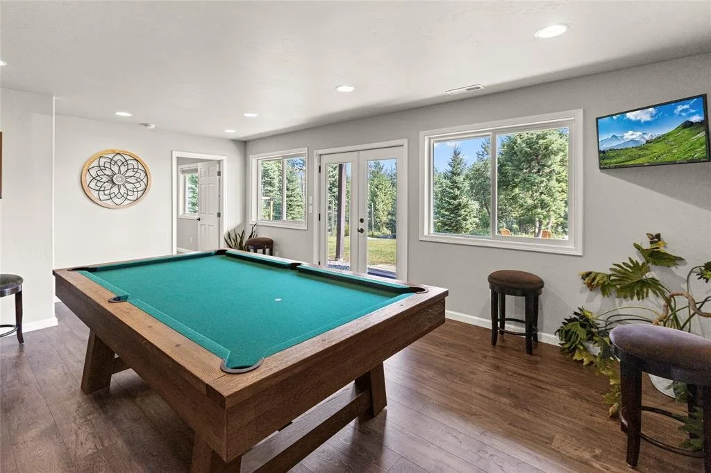 A game room with a pool table, multiple windows looking out on trees, and a wall-mounted TV.