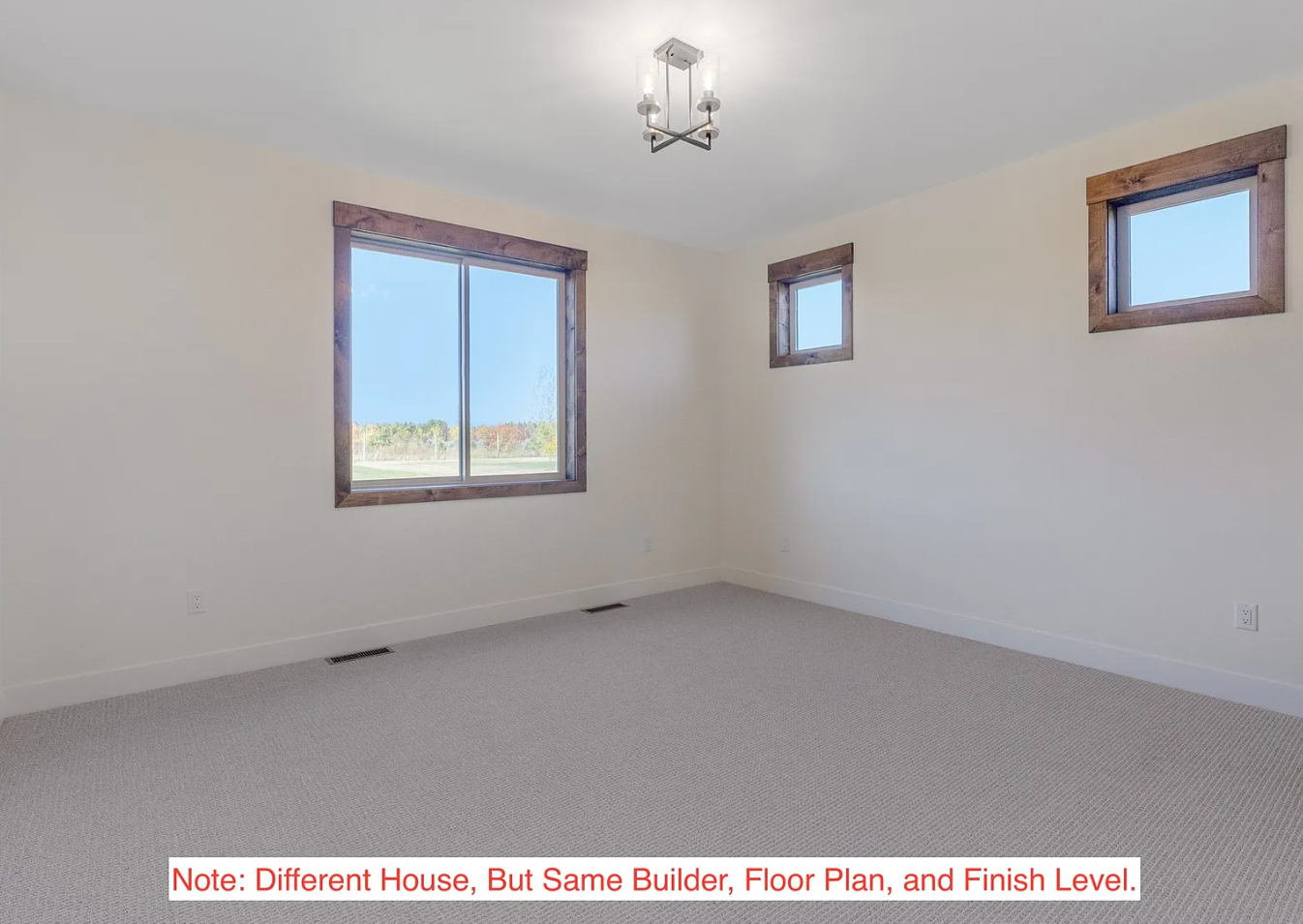 Empty room with beige walls, three windows framed with dark wood, and a modern ceiling light fixture, with light-colored carpeted flooring.