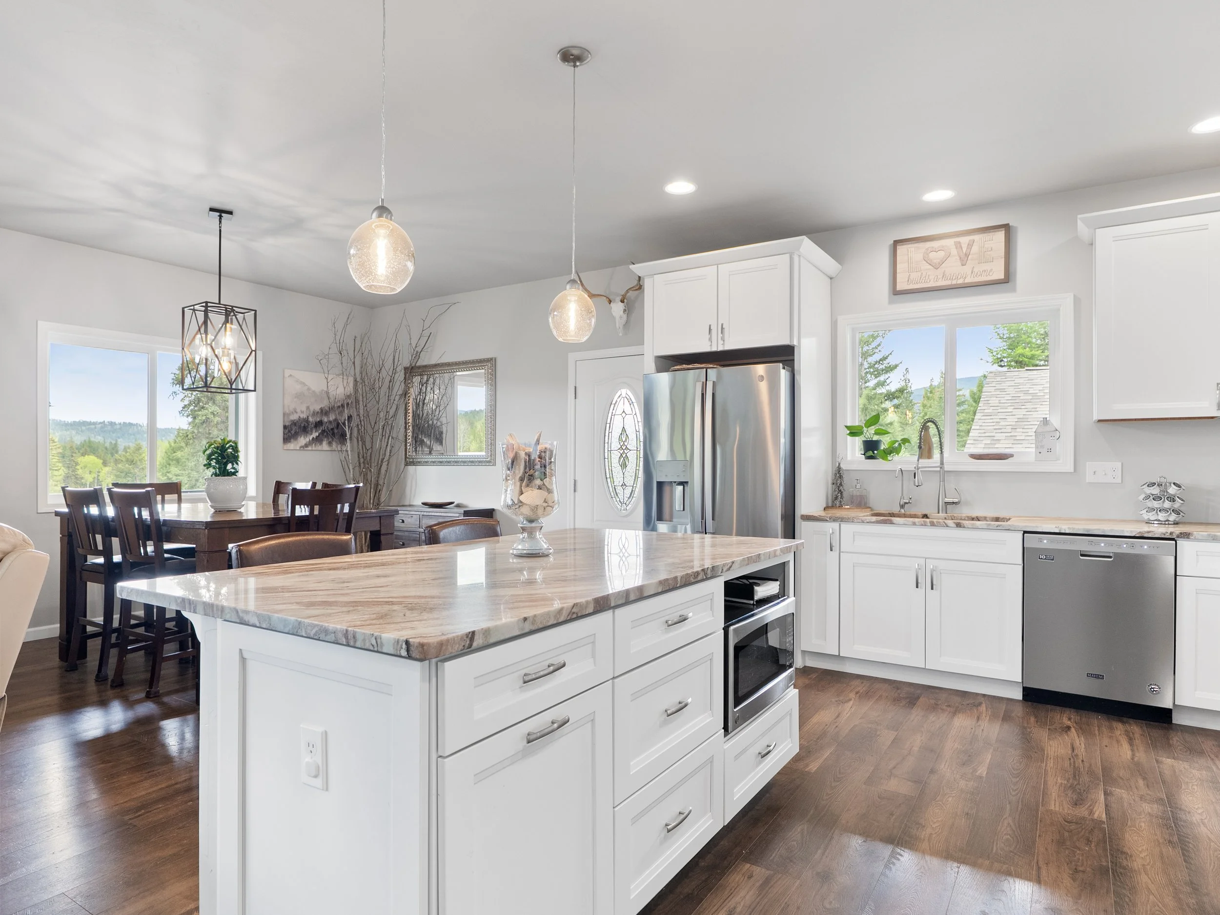 A modern kitchen with white cabinets and a marble countertop island. Stainless steel appliances, including a refrigerator and oven, are visible. Large windows provide natural light and scenic outdoor views. The dining area features a wooden table wit