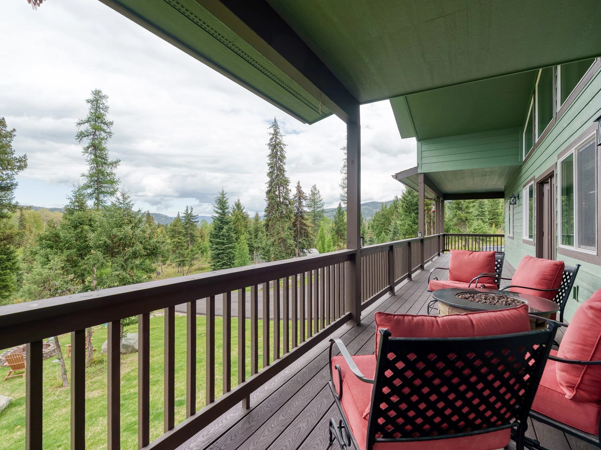 A balcony with green walls and a green ceiling, furnished with red chairs and a round coffee table, overlooking a lush green forest of pine trees.