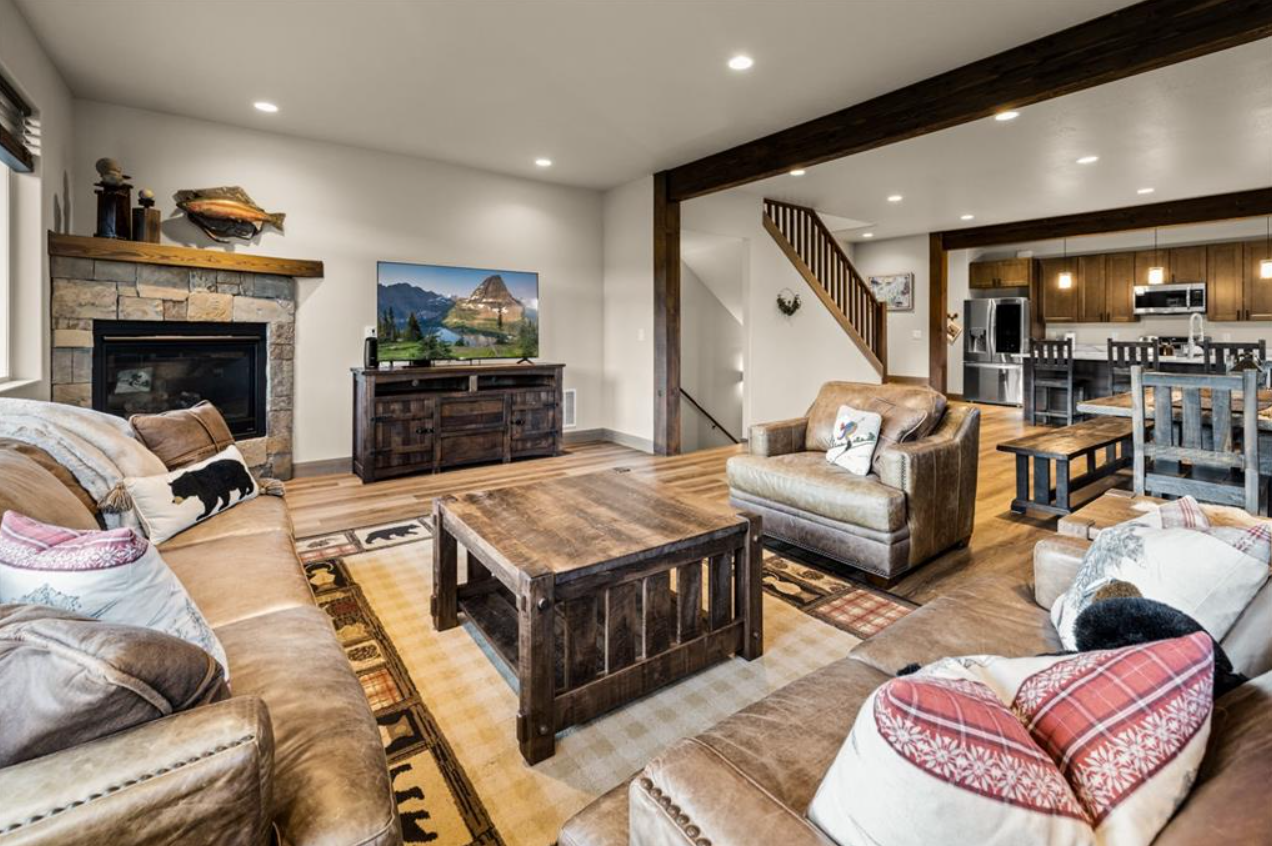 Living room with brown sofas, a stone fireplace, a television on a wooden stand, a wooden coffee table, and view into the kitchen with dining table and chairs, hardwood floors, and wooden beams.