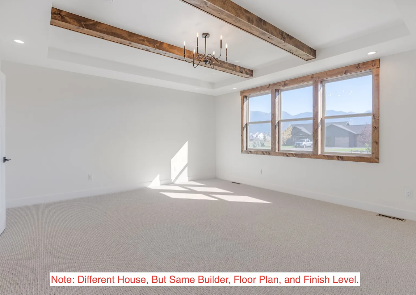 Empty room with white walls, beige carpet, large window with wooden trim, and wooden beams on the ceiling, with mountain view outside.