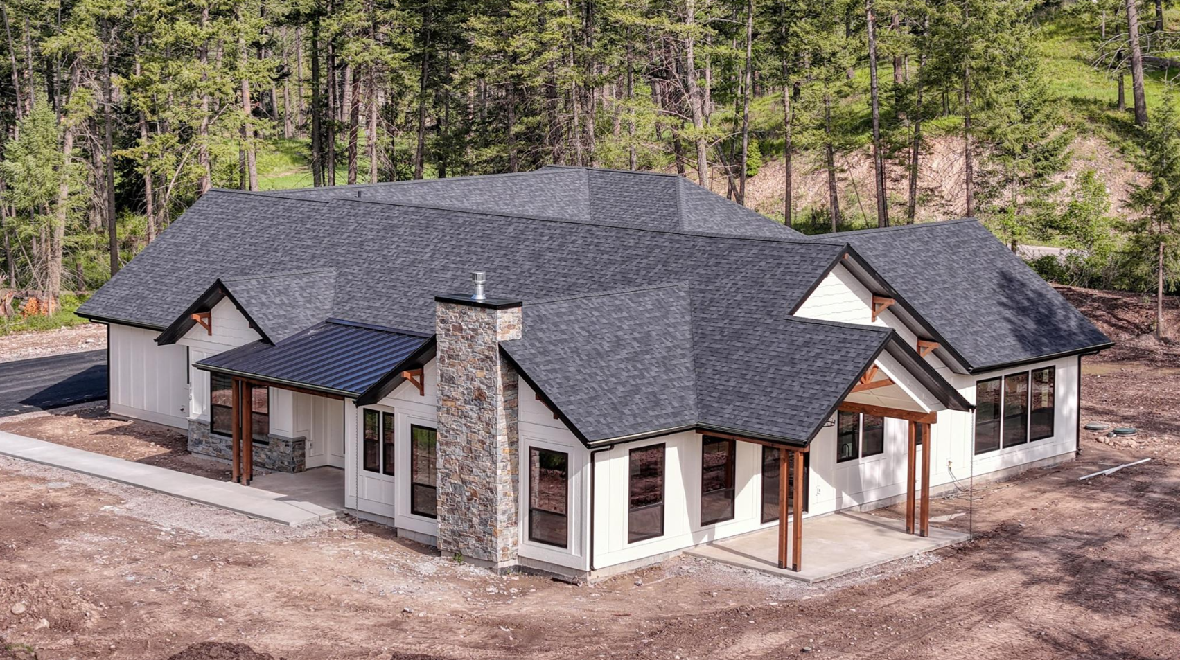 A newly constructed house with a dark shingled roof, light-colored siding, stone chimney, and wooden accents, situated on a partially developed lot with surrounding trees.