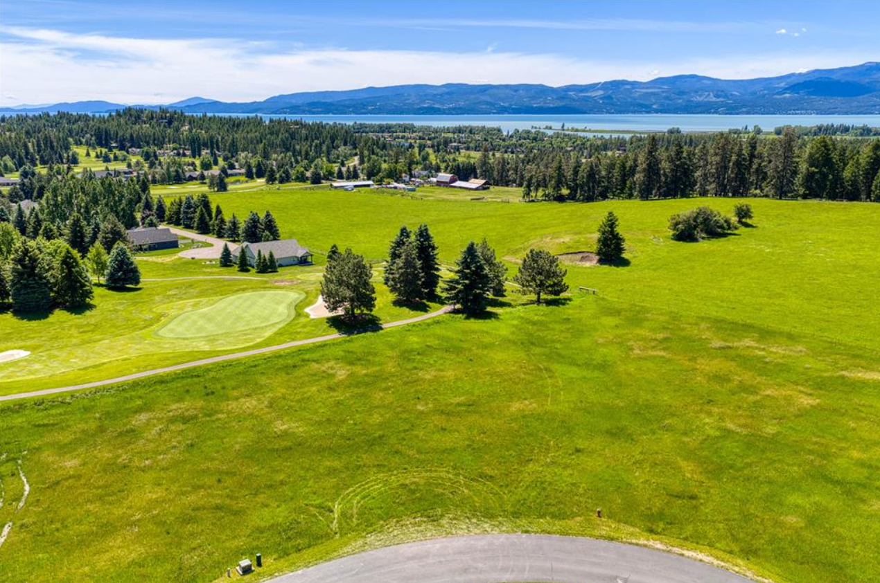 Aerial view of a golf course with green fairways and sand bunkers, surrounded by trees and a few buildings, with a large body of water and mountains in the background under a partly cloudy sky.