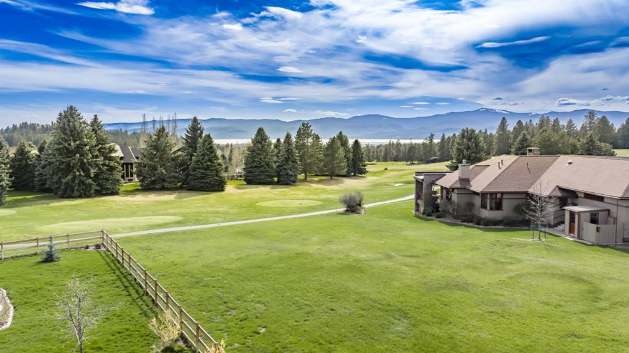 A large grassy yard with a house on the right, trees and a mountain range in the background, and a blue sky with clouds.