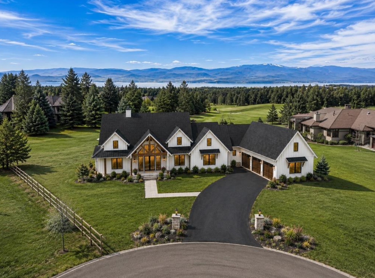 A large white house with a black roof and attached garage, surrounded by green lawns, trees, and a landscaped front yard, with mountains in the background under a partly cloudy sky.
