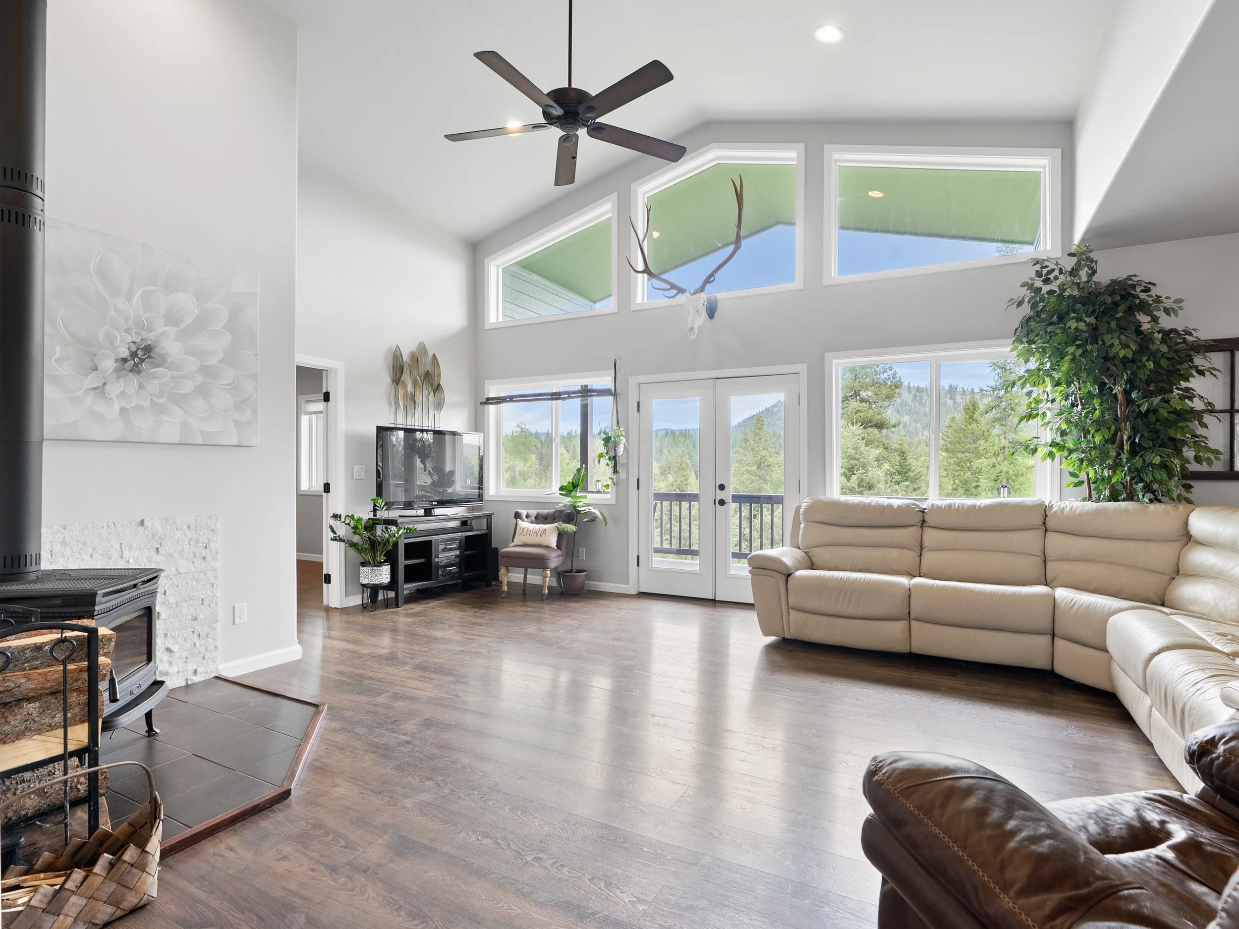 A bright living room features white walls, large windows, a beige sectional sofa, a dark wooden entertainment center with a television, and indoor plants. The ceiling has a black fan, and there's a view of a forest outside.