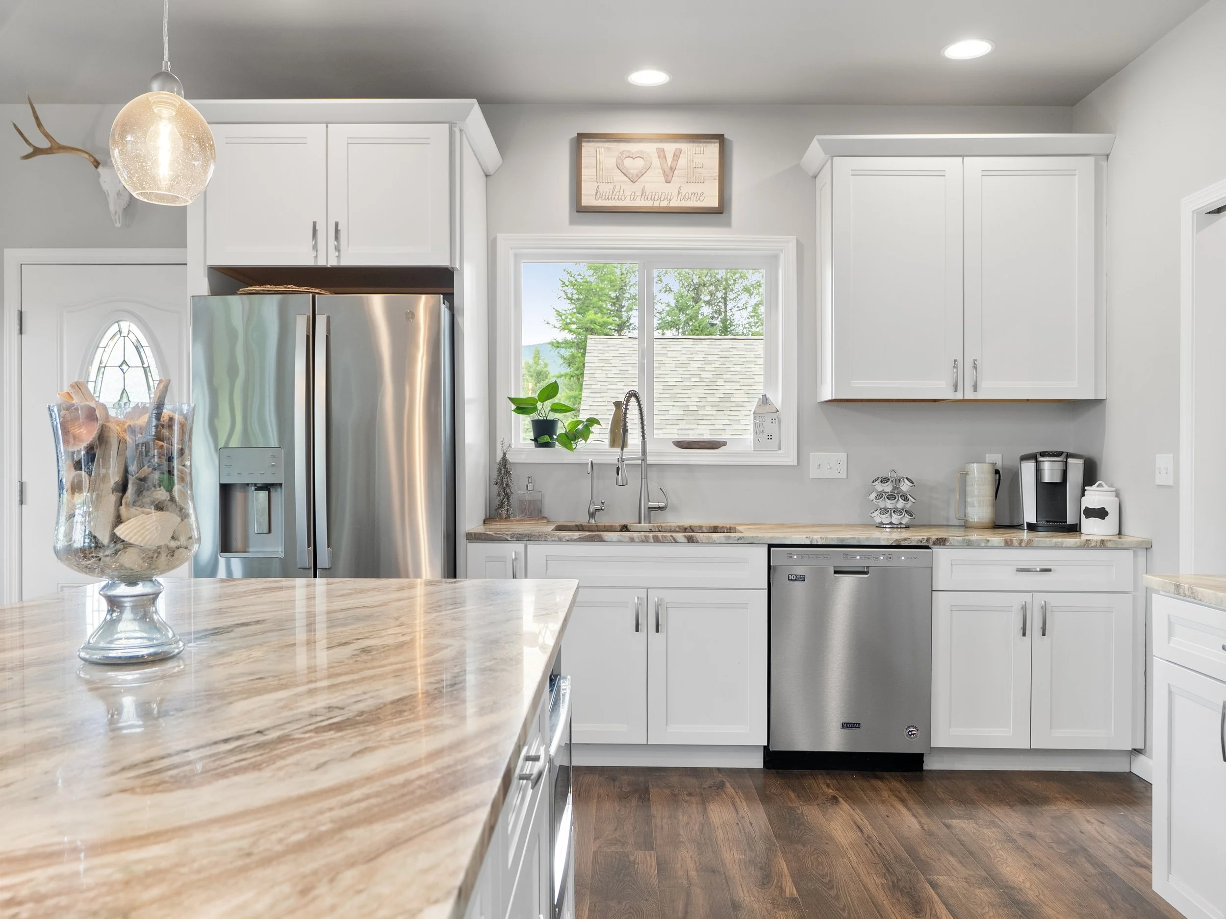 A modern white kitchen with a granite countertop, stainless steel refrigerator, and a window behind the sink showing trees outside. There is a decorative sign above the window that reads 'LOVE builds a happy home' and various small appliances on the 