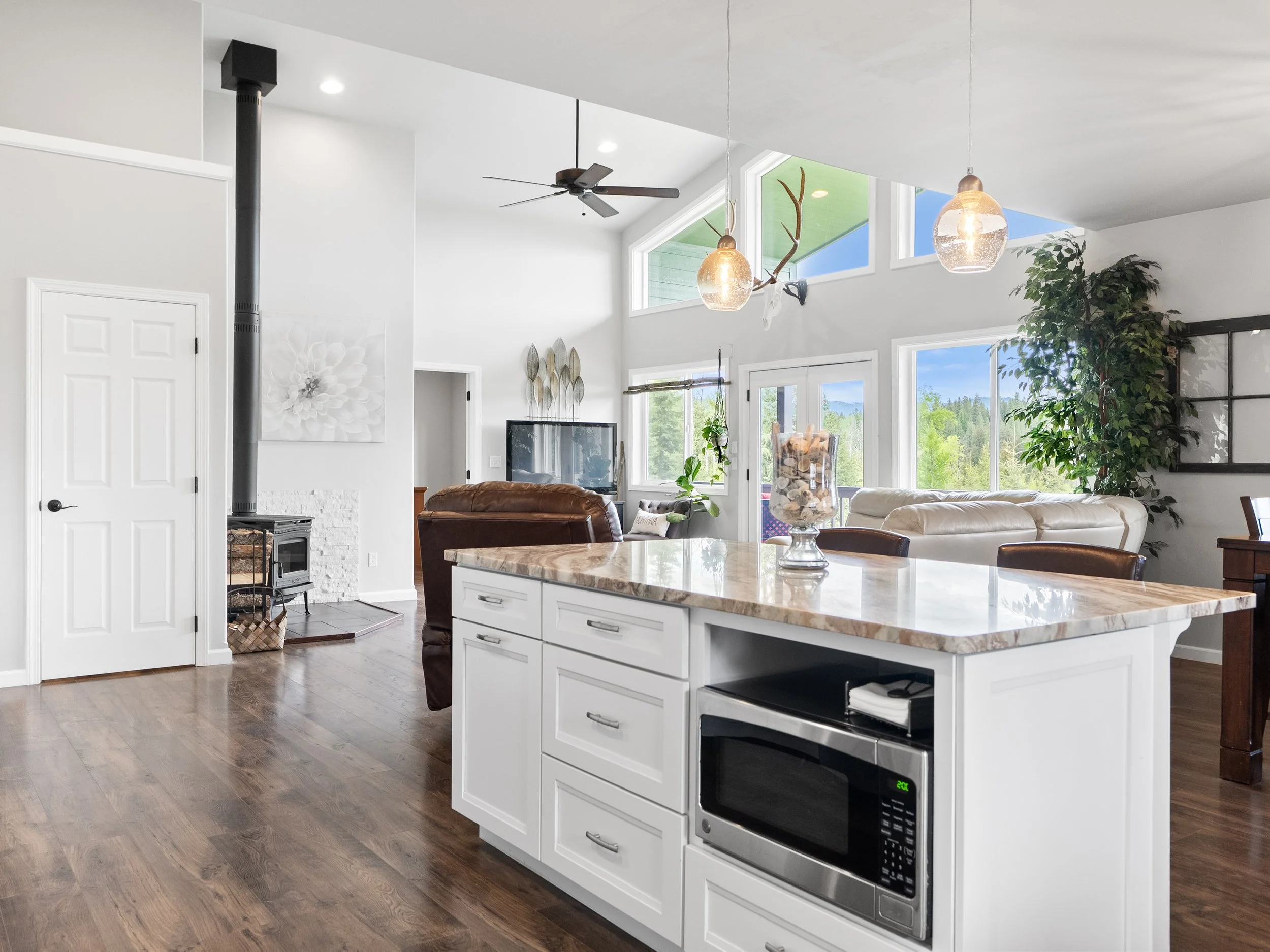 Modern open-concept living space with white kitchen island, brown leather chairs, green plants, large windows with scenic outdoor view, and decorative lighting fixtures.