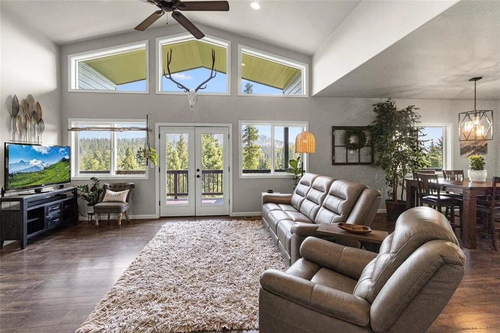 Living room with large windows, a ceiling fan, a TV on a black stand, beige leather armchair and sofa, and a dining table in the background, decorated with plants and a deer skull wall art.